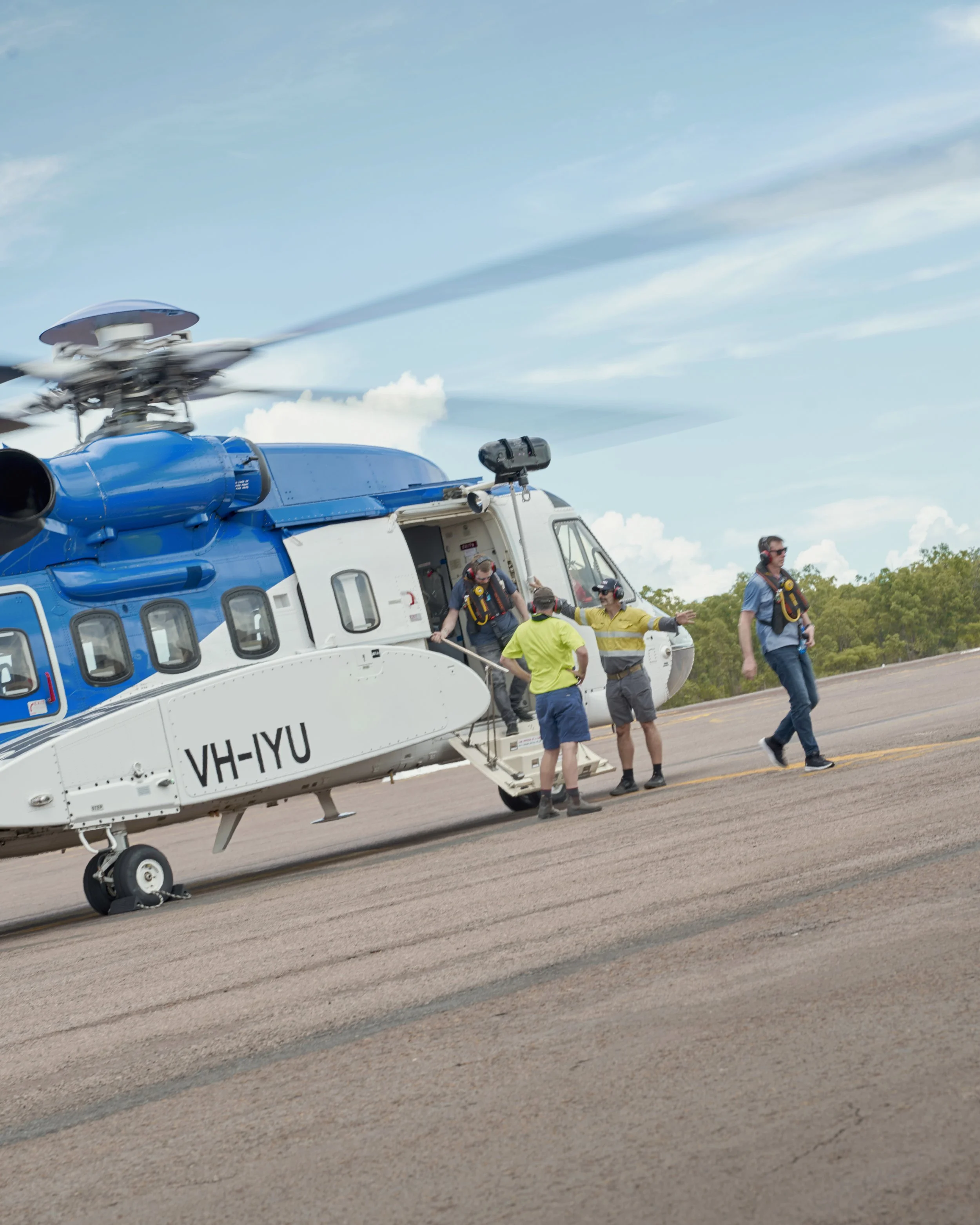 A group of people disembarking from a blue and white helicopter on a tarmac with a clear sky and green trees in the background.