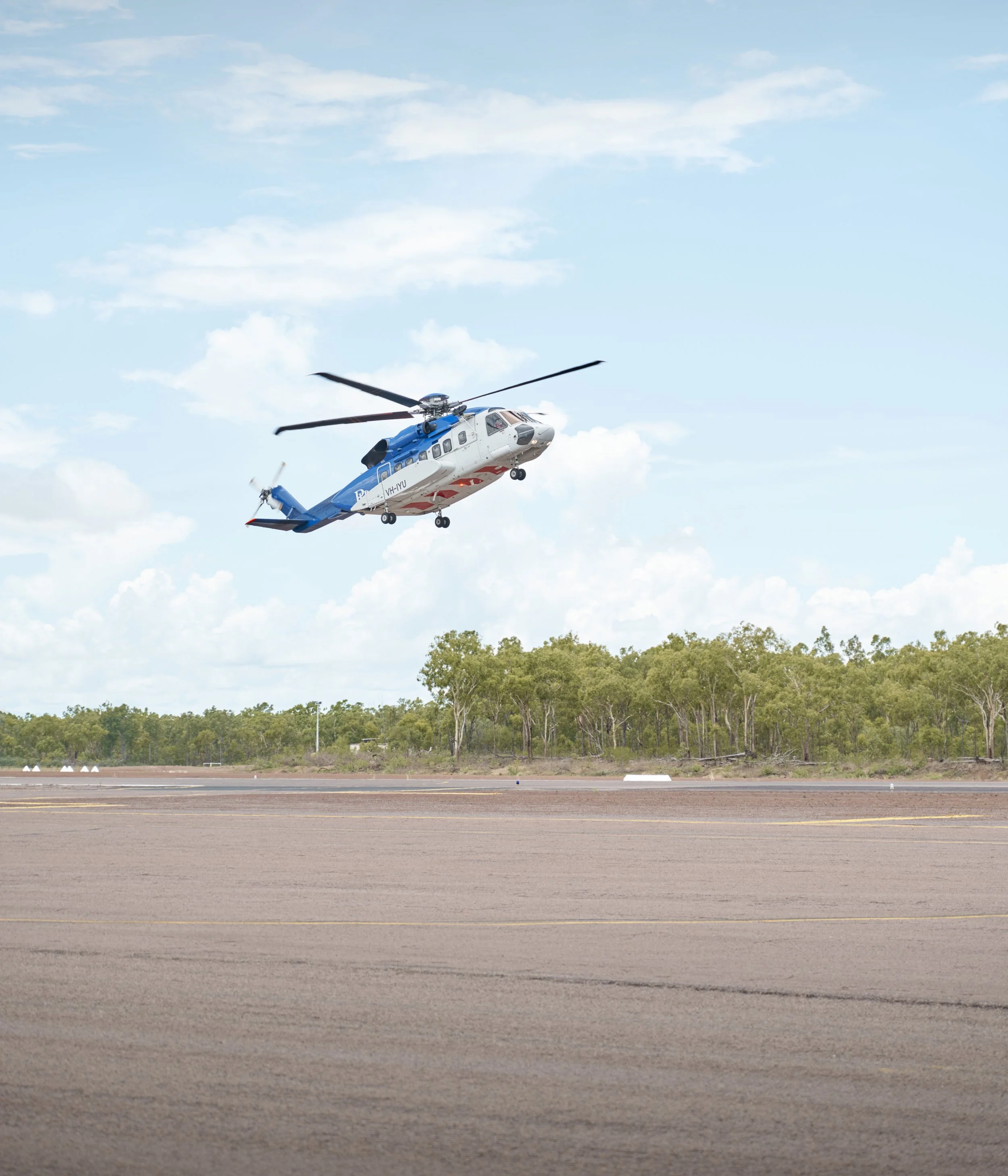 A helicopter flying above a runway with trees in the background and a blue sky with clouds.