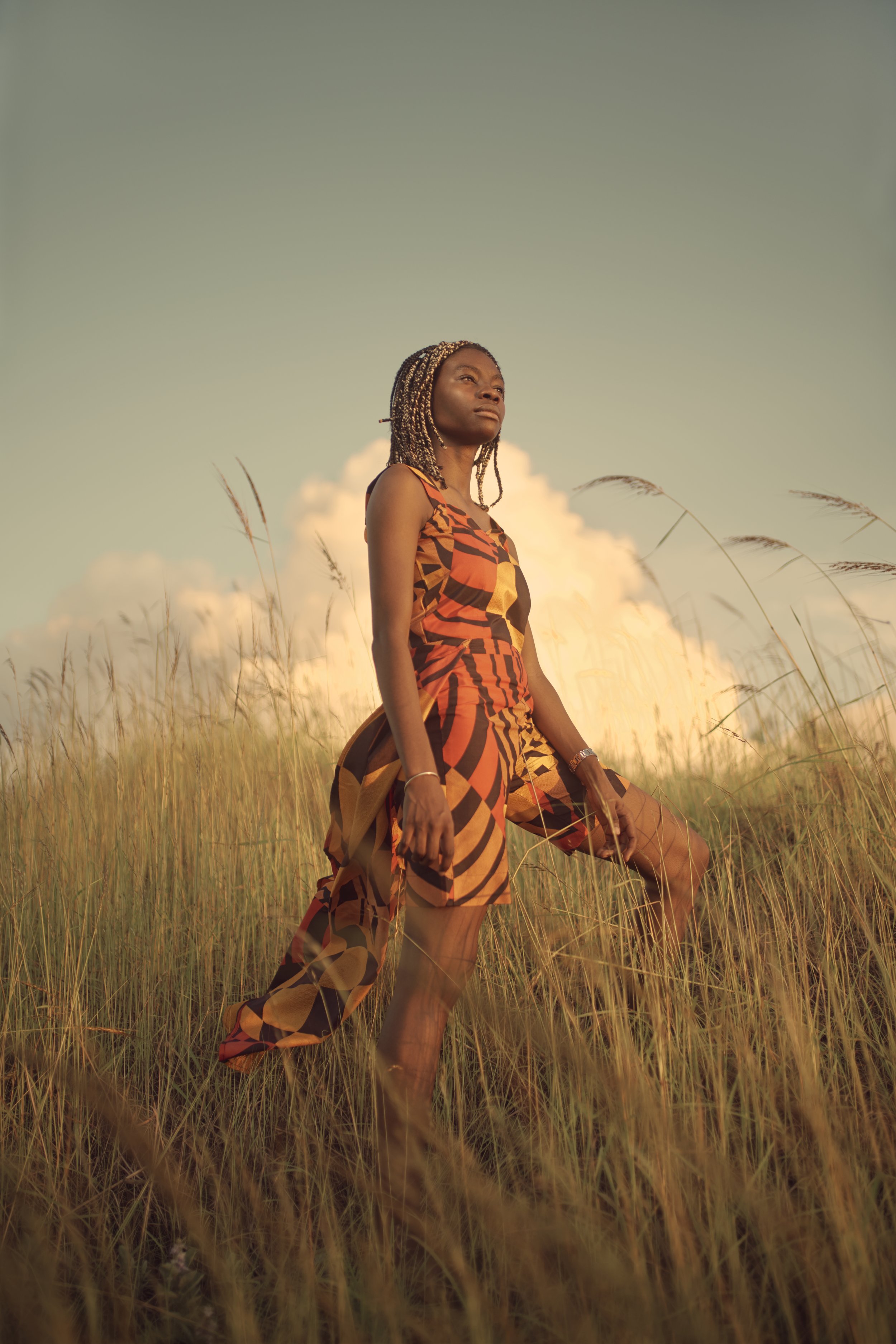 A woman stands in a field of tall grass during golden hour, wearing a colorful patterned dress, with her gaze directed upward and a serene expression.