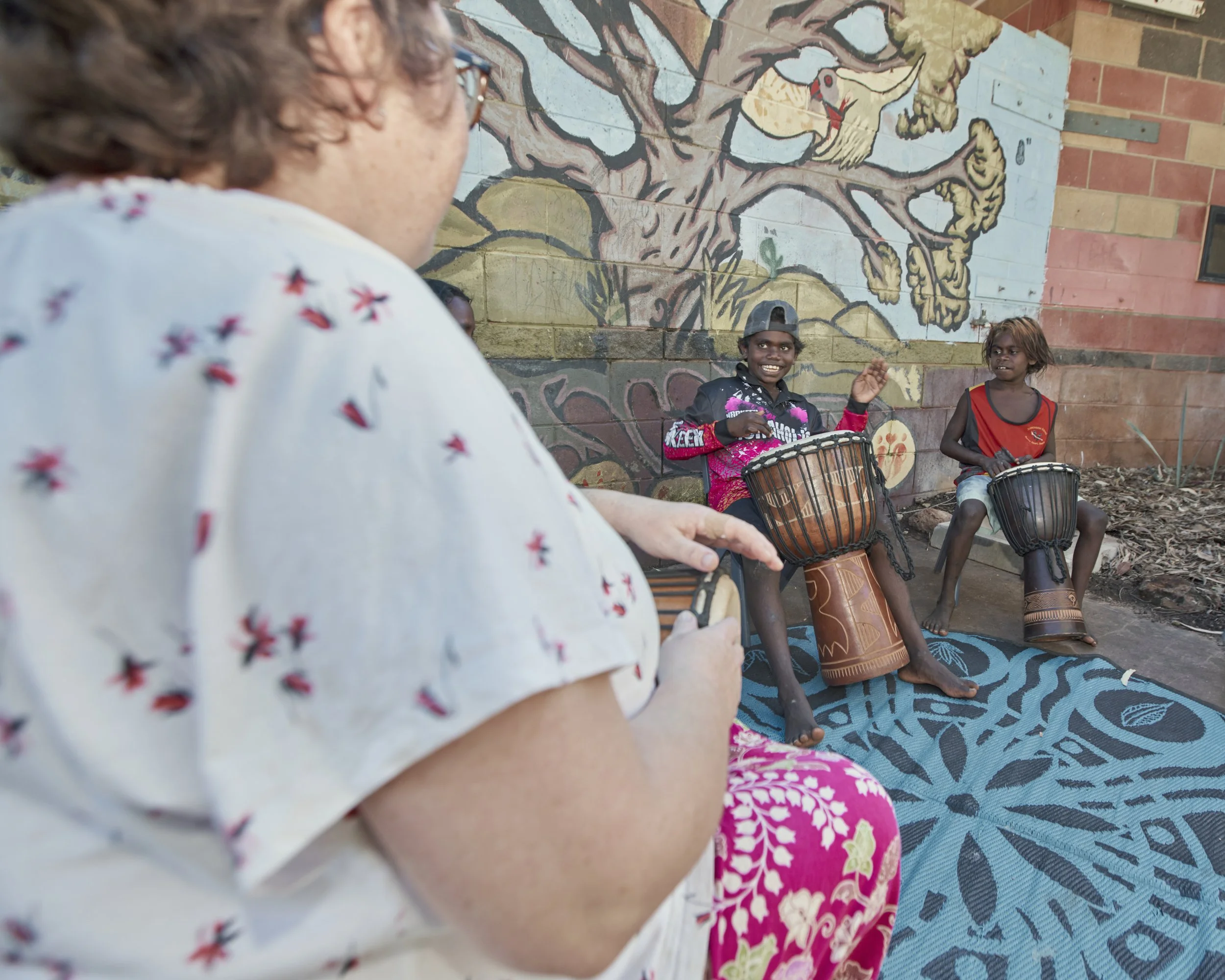 An elderly woman is sitting outdoors, watching two children playing drums against a colorful mural painted on a brick wall. The children are smiling and appear to be enjoying playing percussion instruments. The ground is covered with a blue patterned