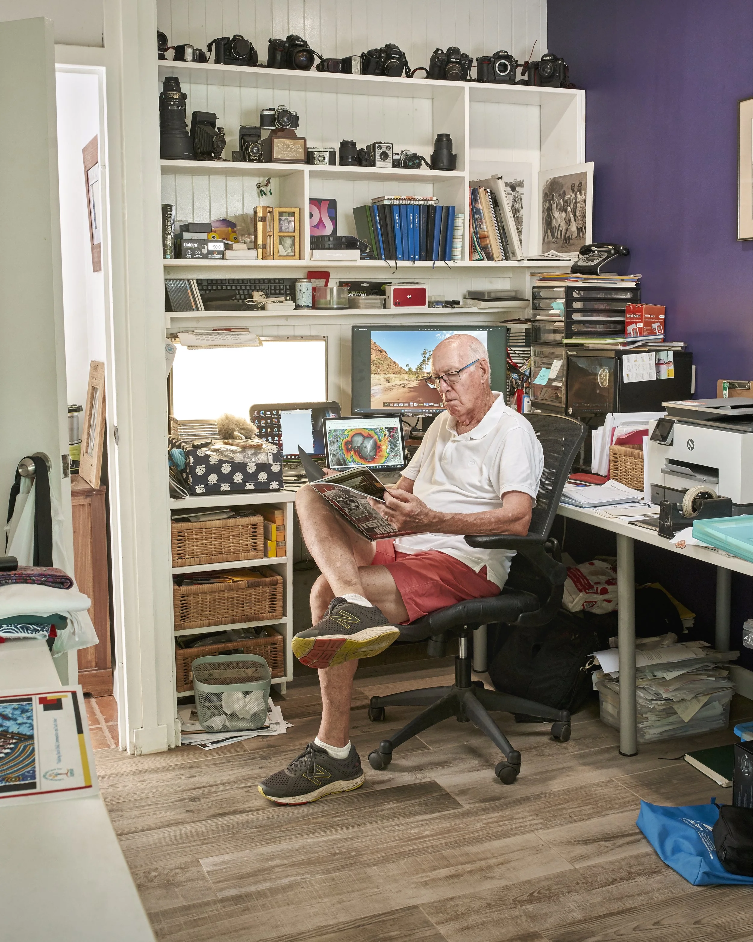 A man sitting in a cluttered home office, wearing a white polo shirt, red shorts, and running shoes, reading a magazine.