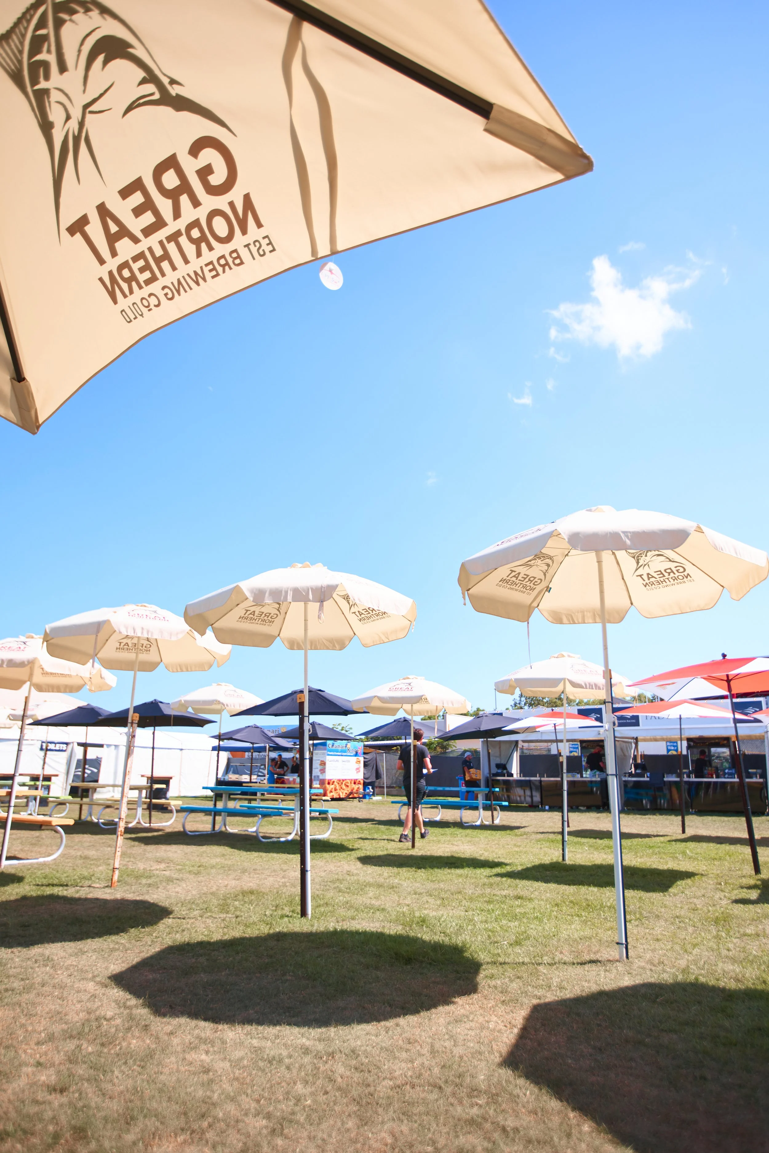 A sunny outdoor scene with multiple white and dark blue umbrellas providing shade over picnic tables on a grassy area at a fair or festival.