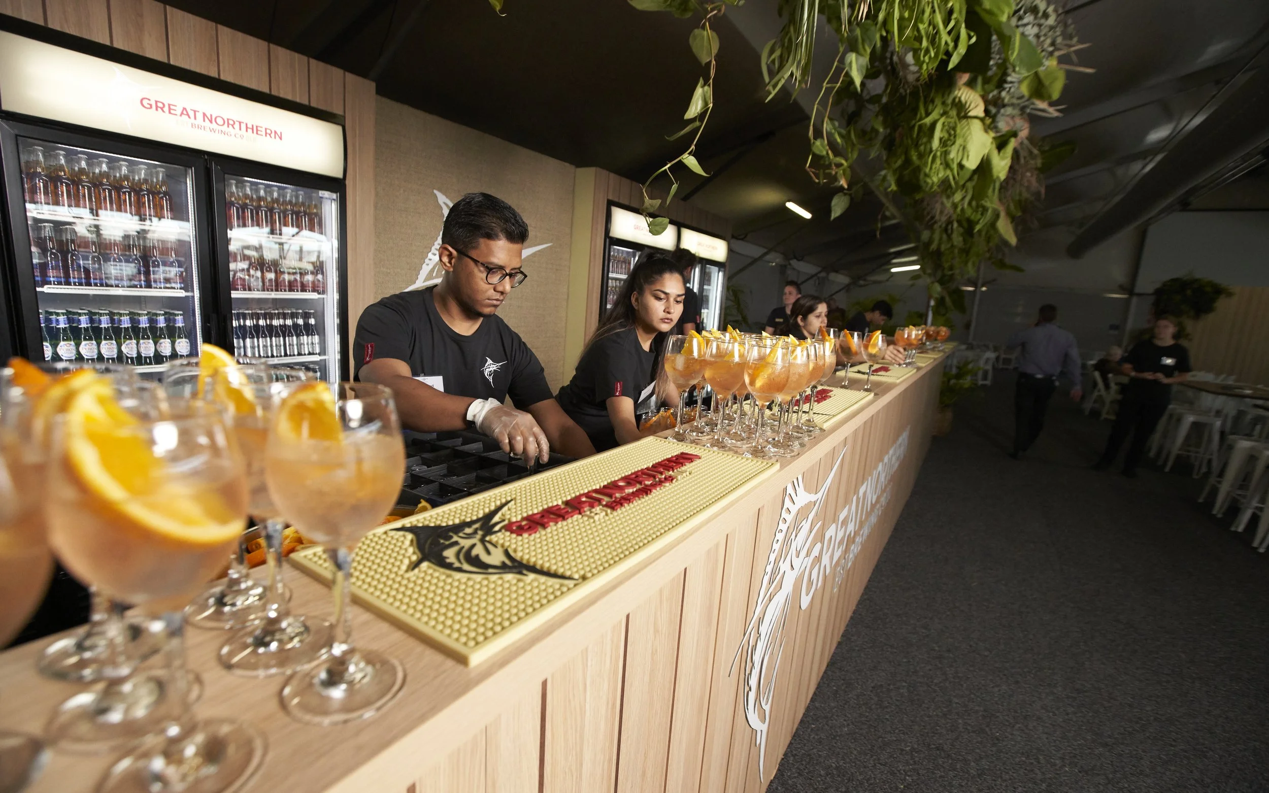 Bar staff preparing drinks at a bar with multiple glasses of orange beverages garnished with lemon slices in front of refrigerated bottles, in a dimly lit event space with hanging green plants.