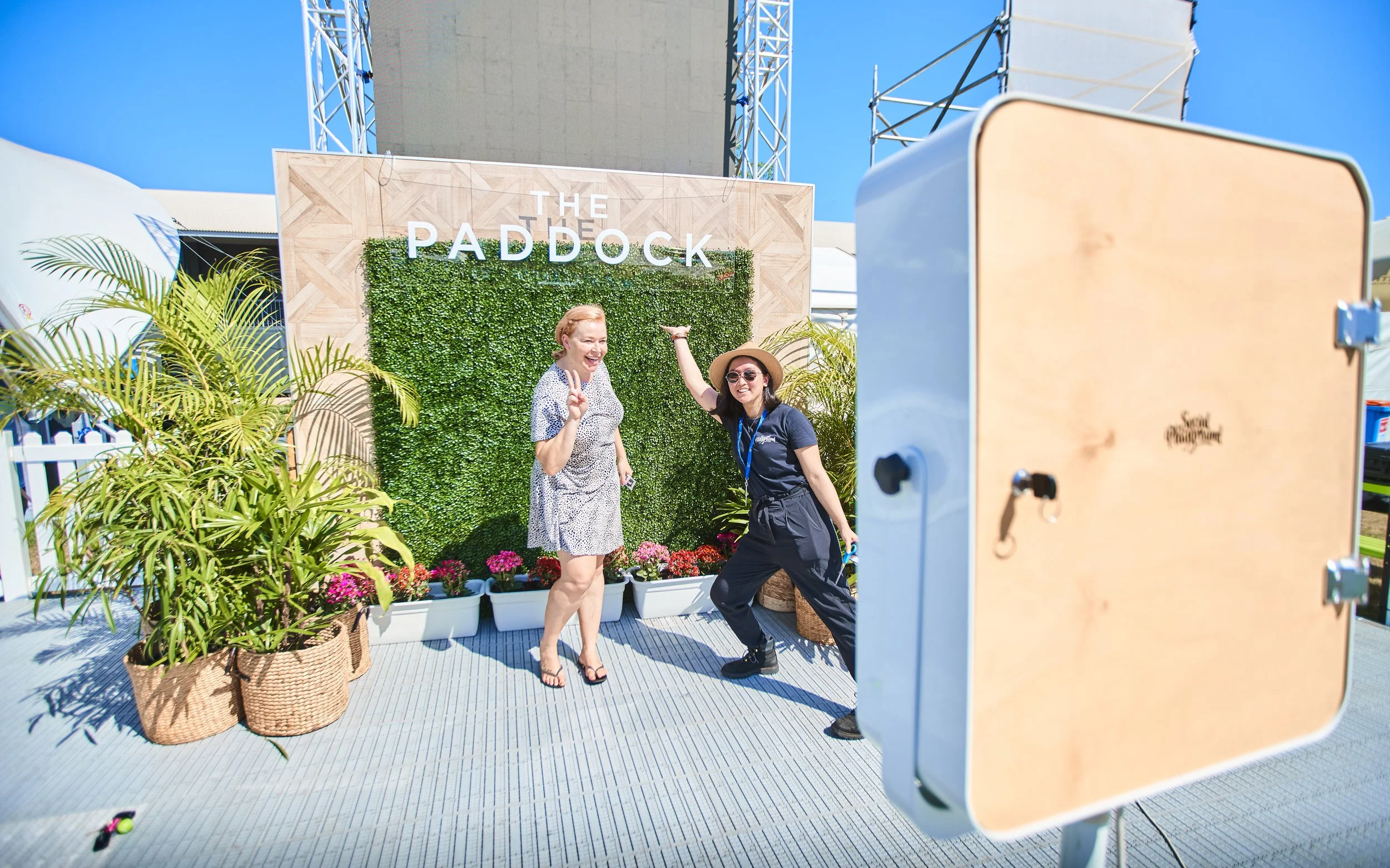 Two women posing and smiling for a photo at an outdoor event called The Paddock, with a green wall and colorful flowers behind them, and a large photo booth in the foreground.