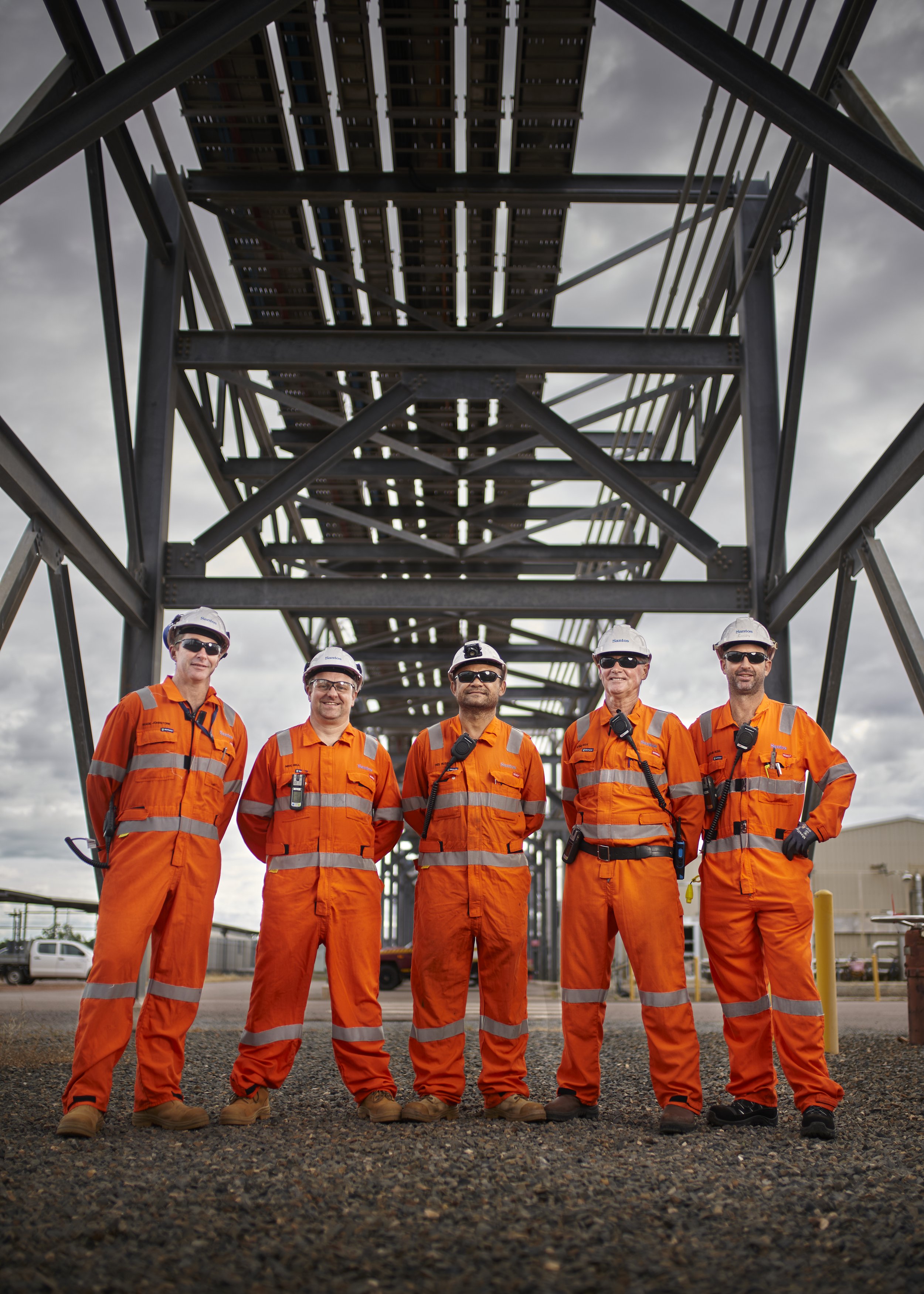 Group of five construction workers in orange safety uniforms and white helmets standing under a large steel structure at an industrial site.
