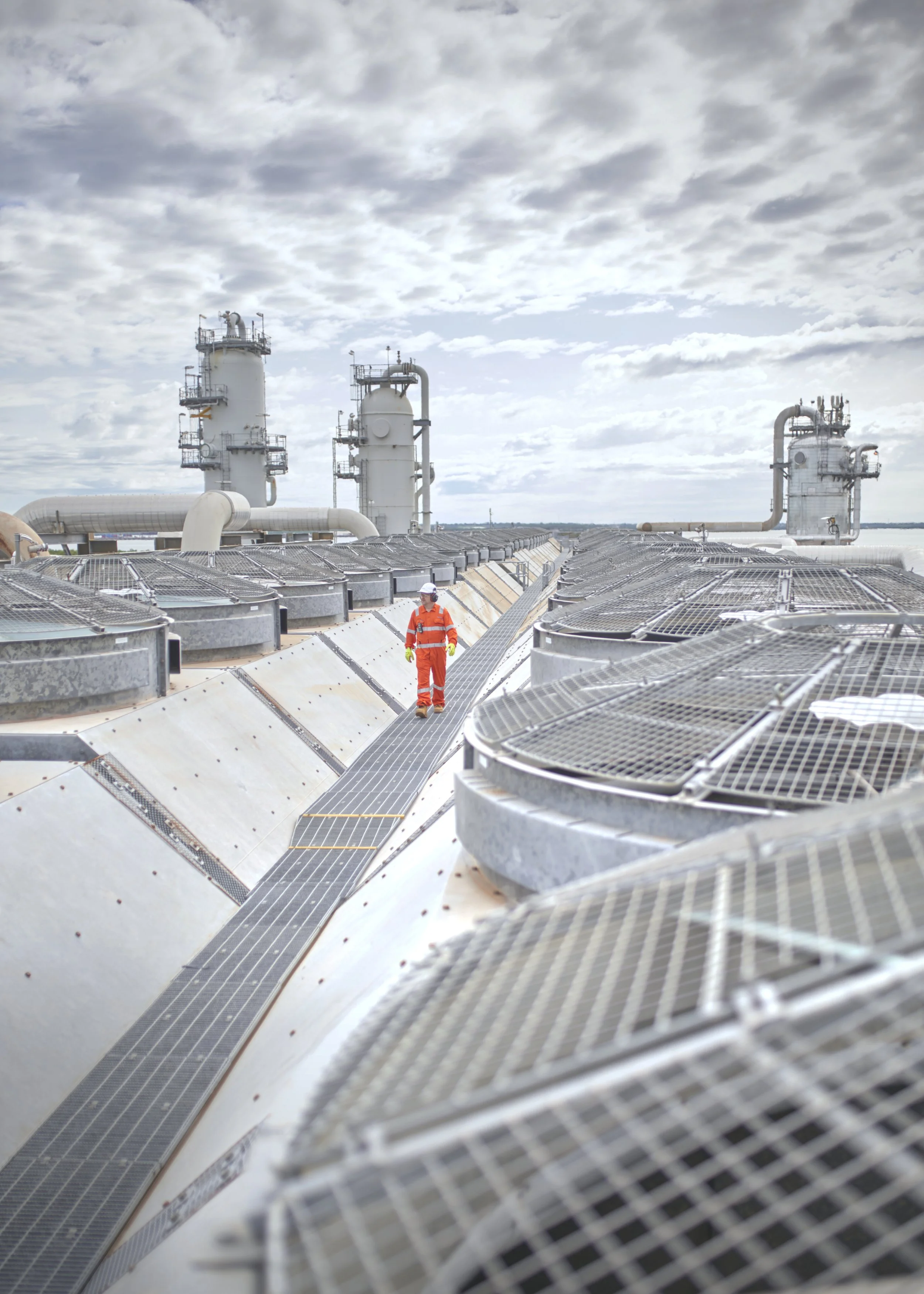 A worker in orange safety gear walking on a large, industrial cooling tower structure with multiple vents and pipes, under a cloudy sky.