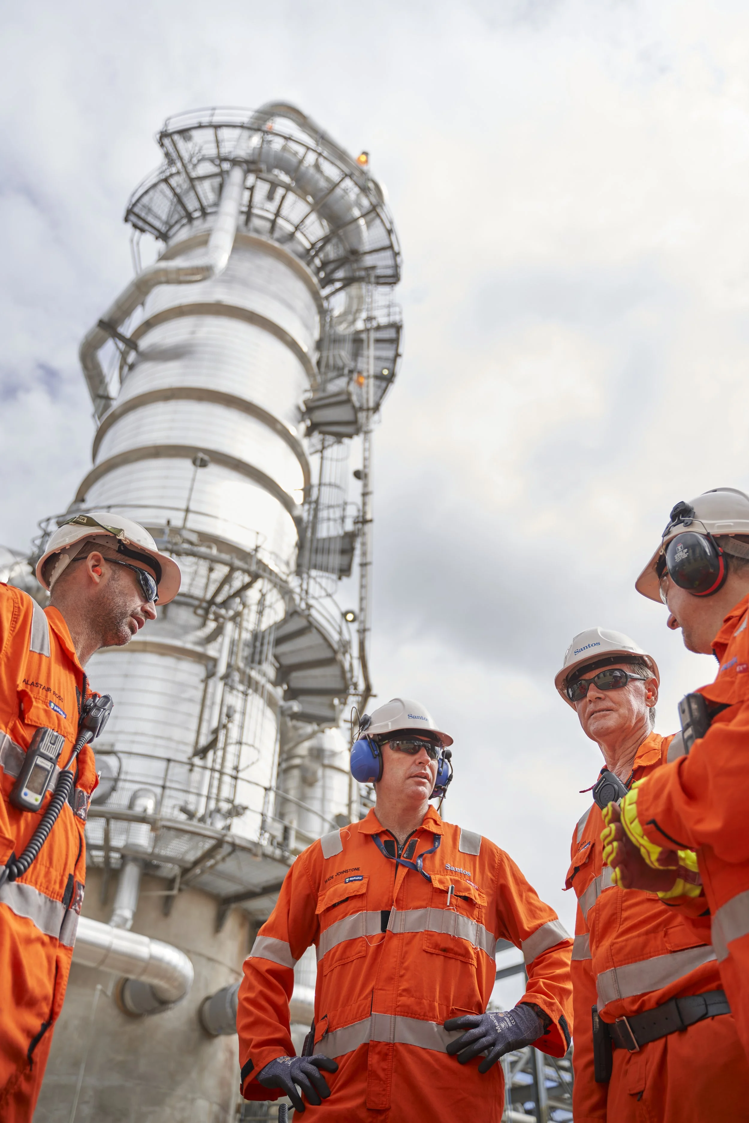 Four workers in orange safety uniforms and hard hats stand in discussion outside an industrial facility with a tall, metallic structure in the background.