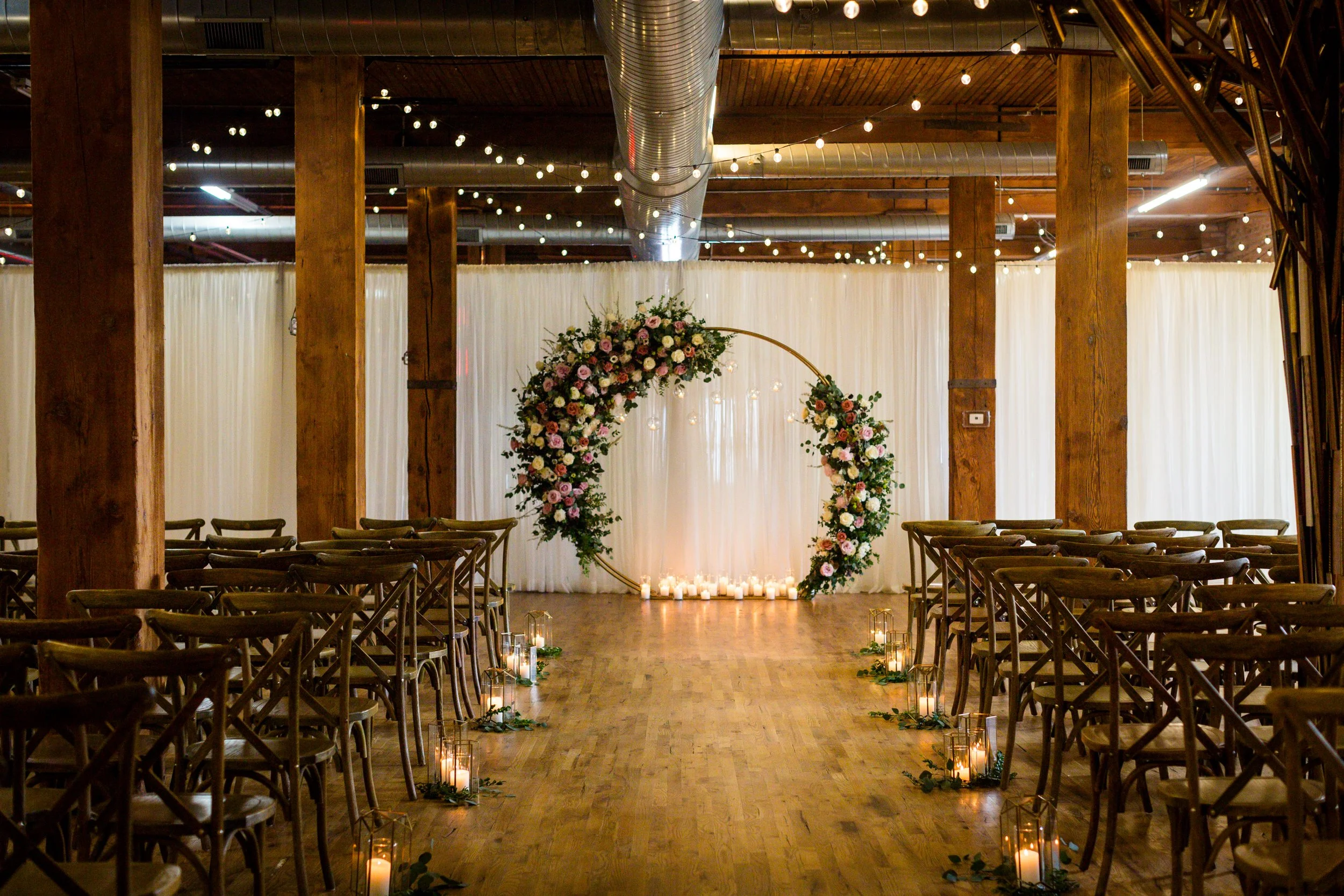 Indoor Lacuna Loft wedding ceremony setup with chairs on each side of a wooden aisle, floral arch at the front, candles along the aisle, string lights overhead, and a white backdrop.