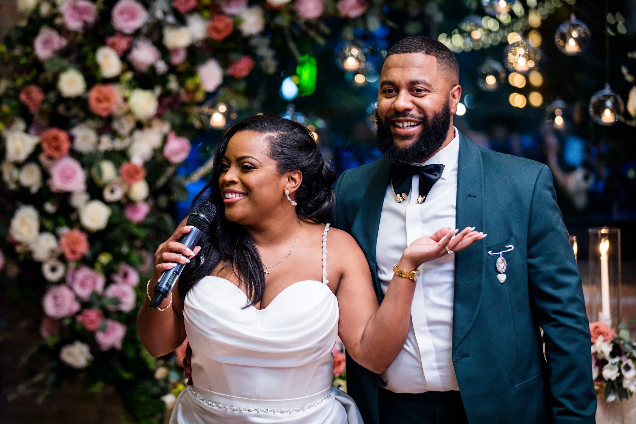 A woman in a white wedding dress holding a microphone and smiling, standing next to a man in a dark green suit with a bowtie, at a wedding reception with floral decorations and hanging lights in the background.