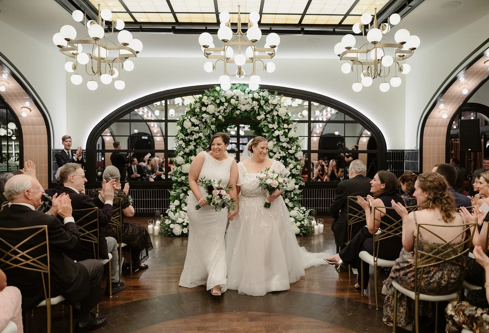 Two brides in wedding dresses holding hands, at the Wellsley Chicago walking down the aisle at a wedding ceremony with guests seated on either side, white floral arch in the background, and modern chandeliers hanging from the ceiling.