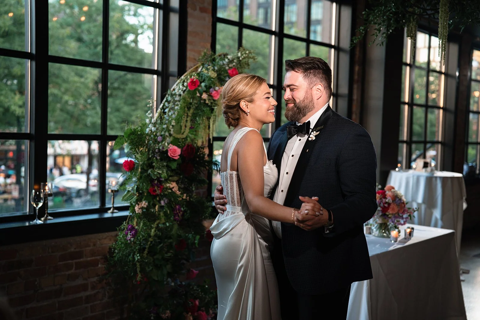 A newlywed couple dancing at their wedding reception indoors with large windows, brick walls, and a floral arch behind them.