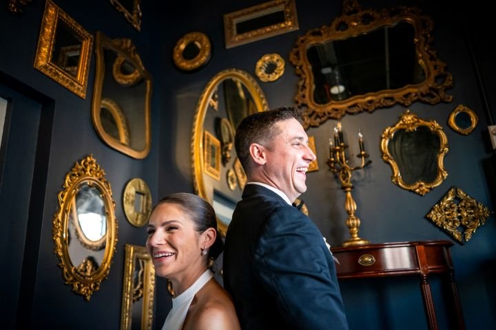 A man and woman smiling and laughing in front of a wall decorated with various gold-framed mirrors and picture frames.