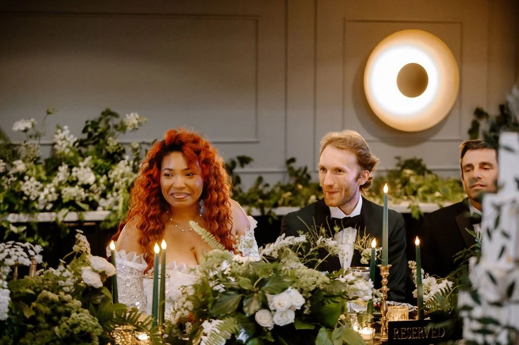 People sitting at a decorated wedding reception table, with floral arrangements, candles, and reserved sign, in a decorated indoor venue.