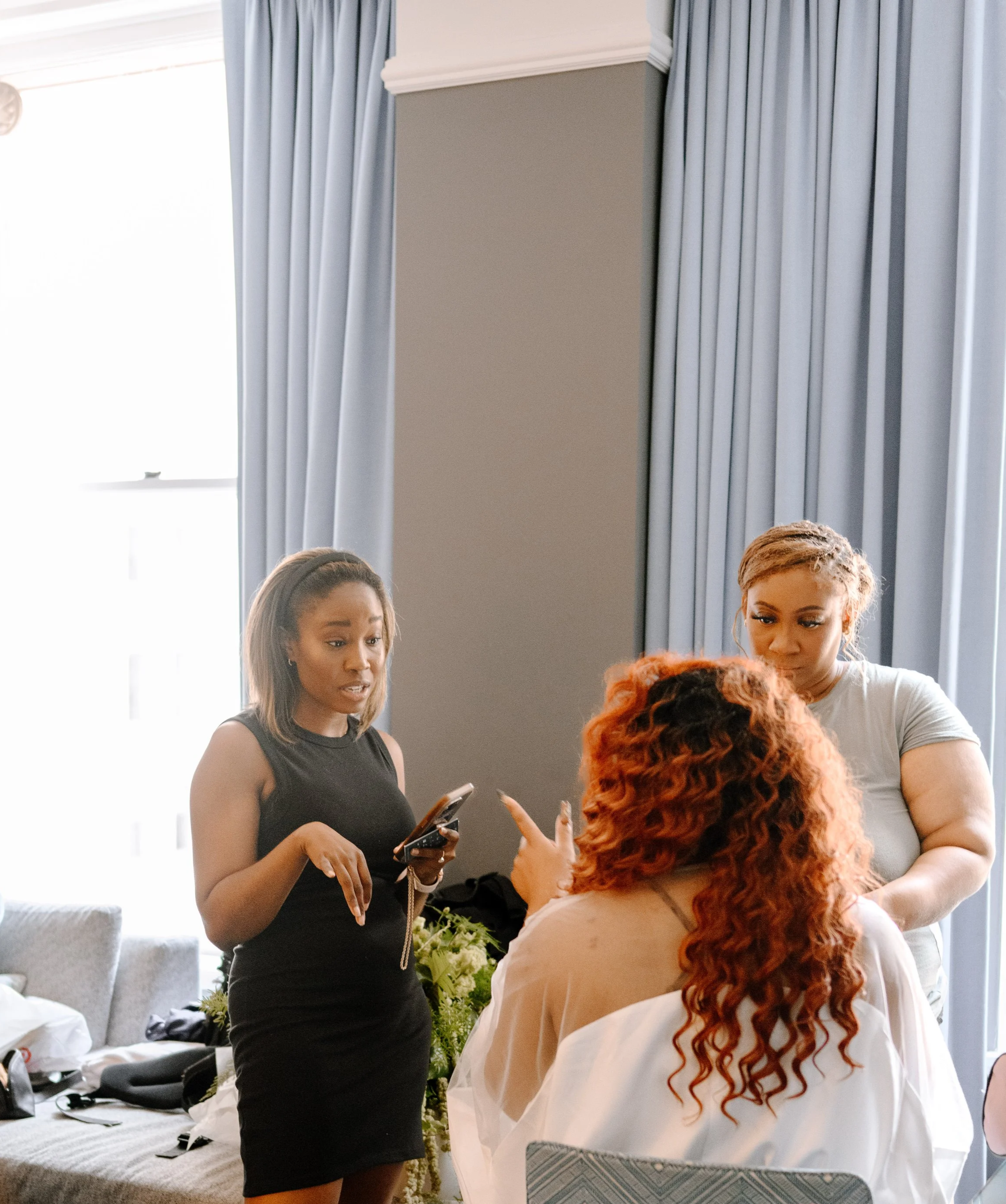 Three women engaged in conversation in a well-lit room with blue curtains; one woman with red curly hair seen from behind, another woman with short brown hair looking at her phone, and a third woman with light brown hair looking at the woman with curly hair.