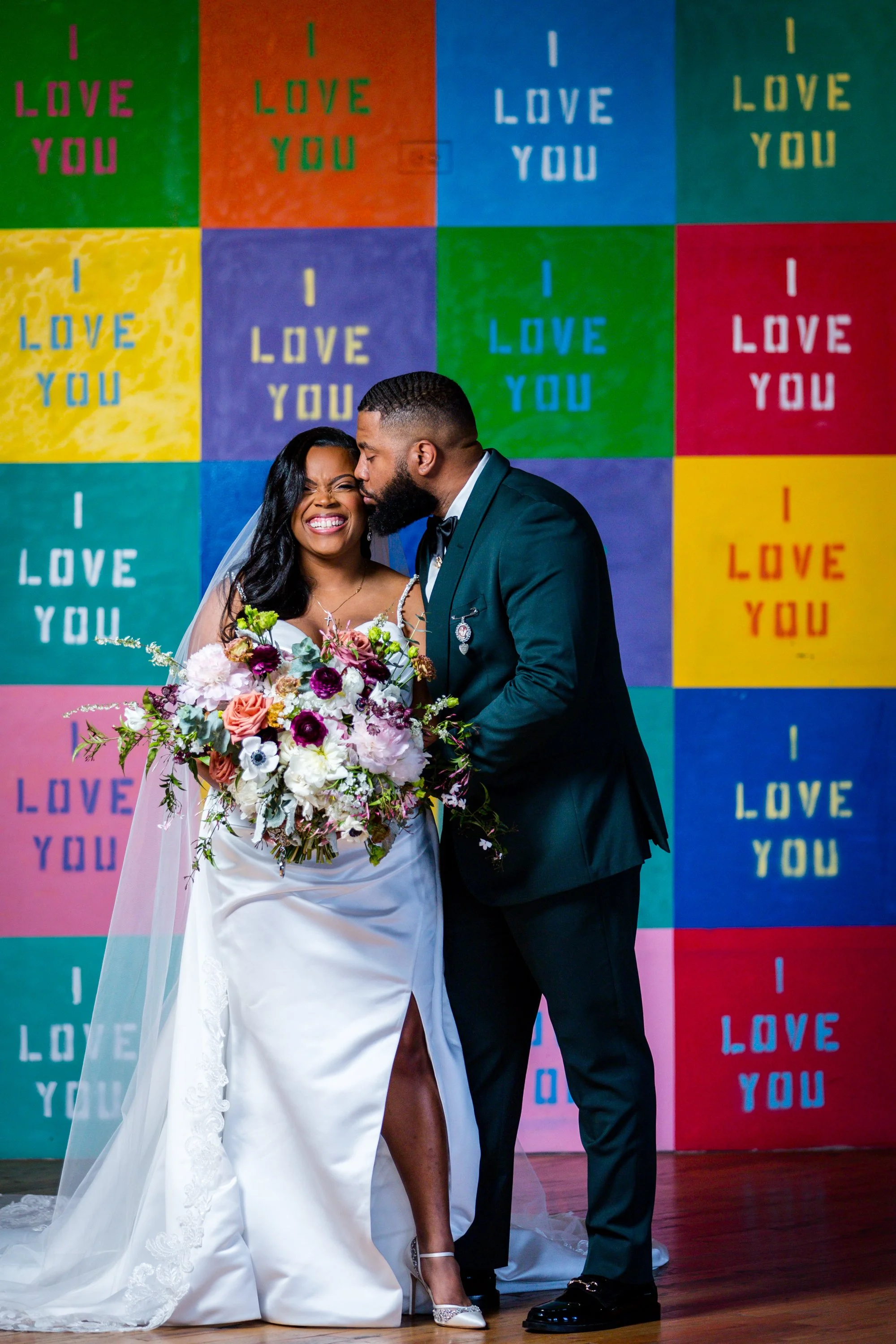 Bride and groom smiling in front of colorful mural that says "I Love You"