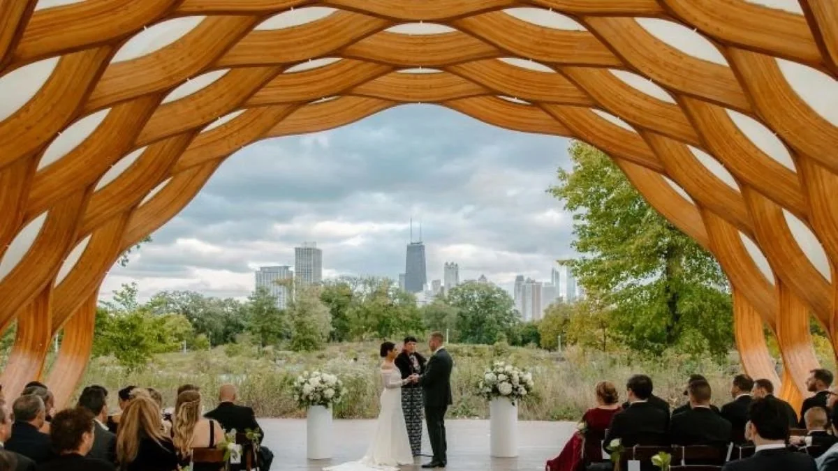 A wedding ceremony taking place outdoors under a large wooden arch with a city skyline in the background. The bride and groom stand facing each other, holding hands, with an officiant between them. Guests are seated on either side, watching the ceremony.