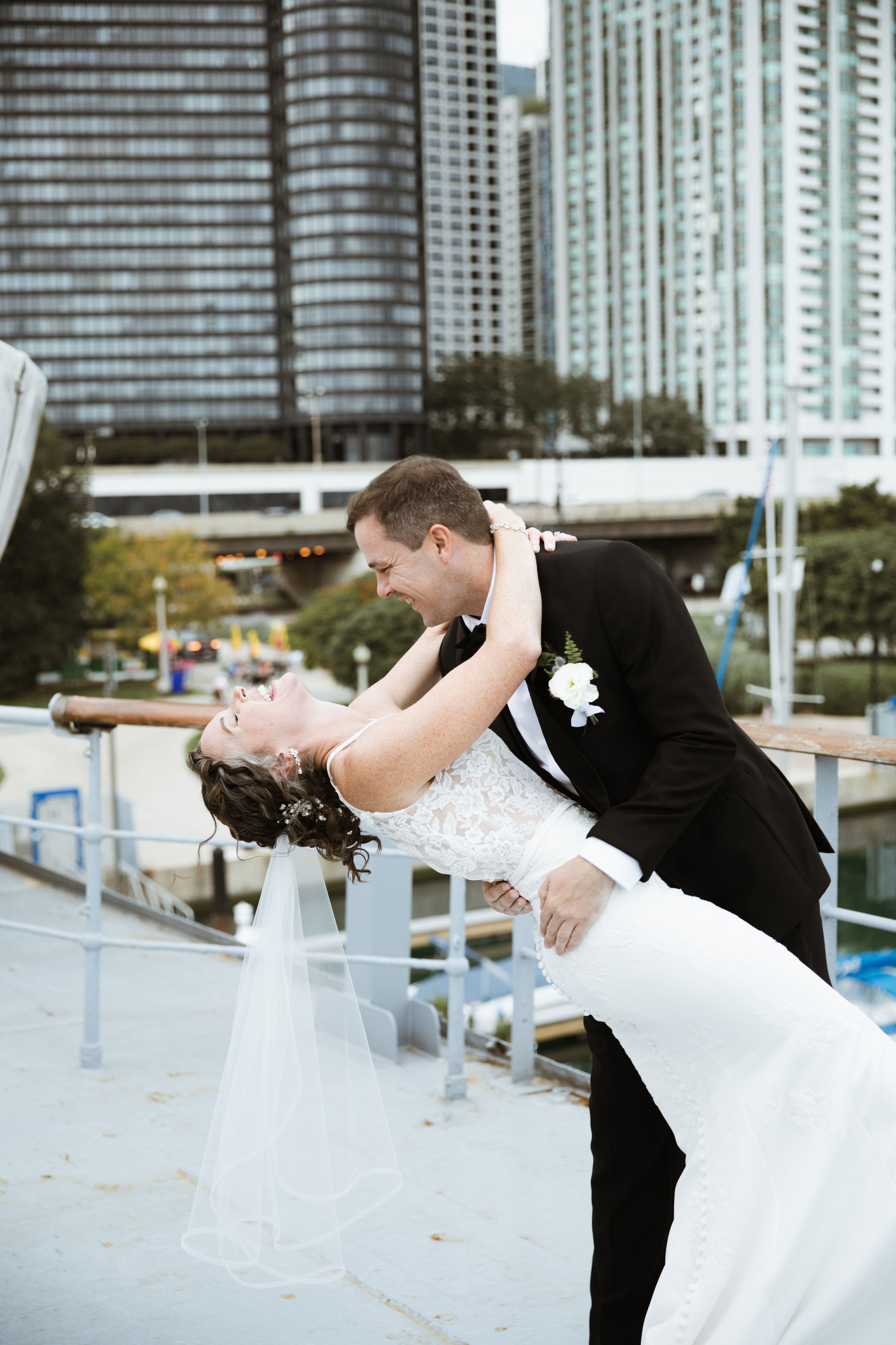 A newly married couple dancing happily outdoors on a city waterfront with tall modern buildings in the background.