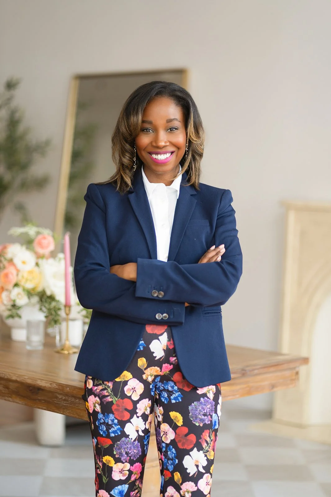 A woman in a navy blazer, white shirt, and floral pants standing with her arms crossed and smiling in a home or office setting with a table, floral arrangement, and mirror in the background.