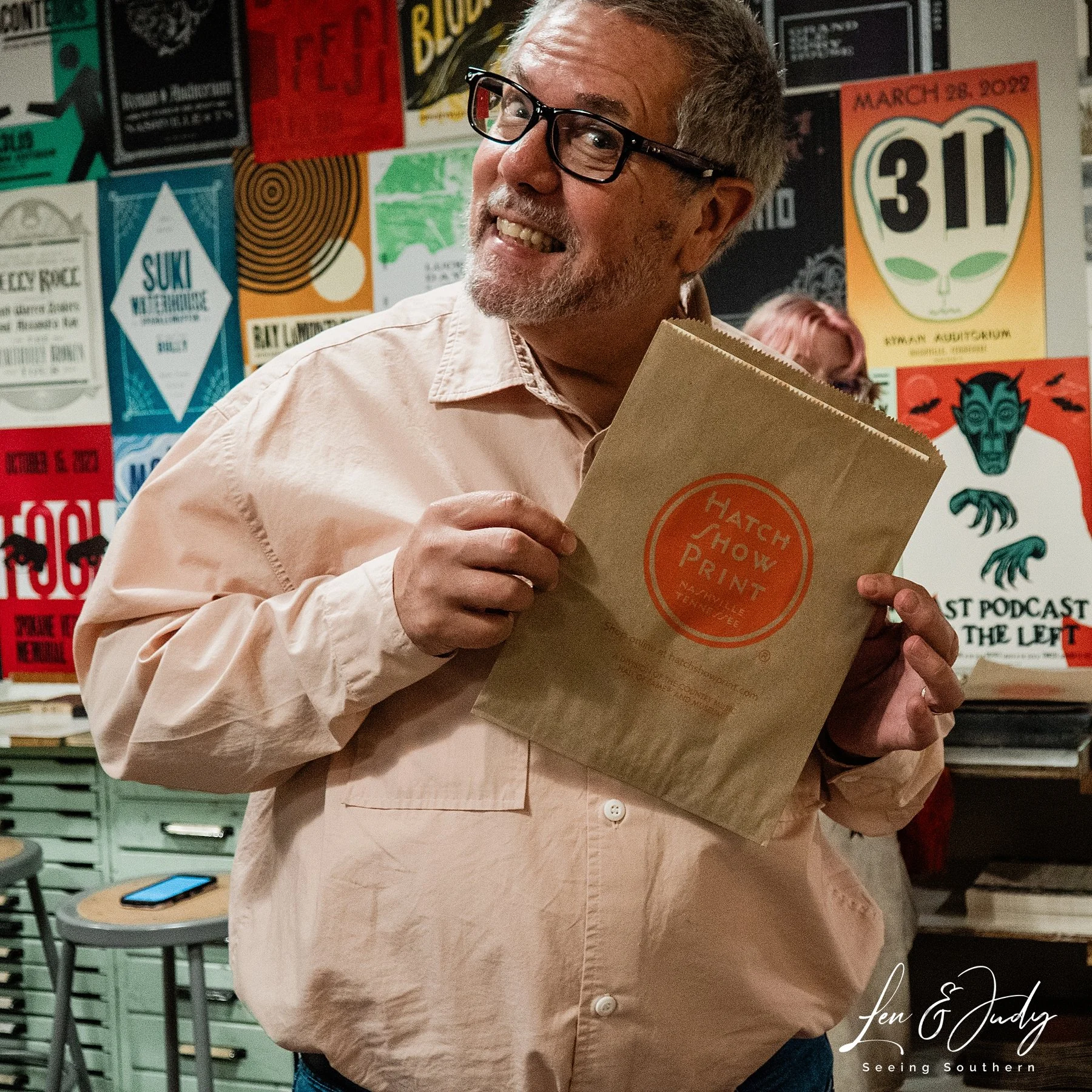 A man with gray hair, glasses, and a beard smiling while holding a paper bag labeled 'Hatch Show Print' in front of a colorful wall covered with various posters.