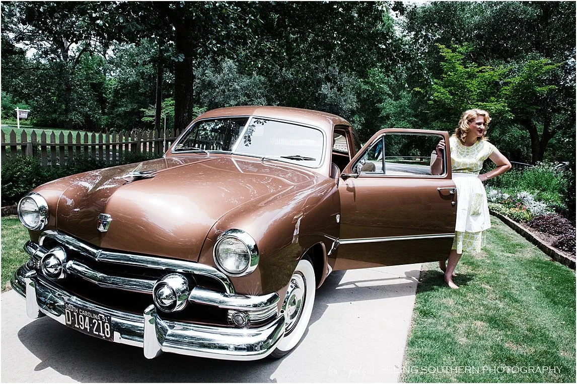 A woman in a yellow and white dress standing beside a vintage brown car with a South Carolina license plate, parked on a sidewalk next to a garden and trees.