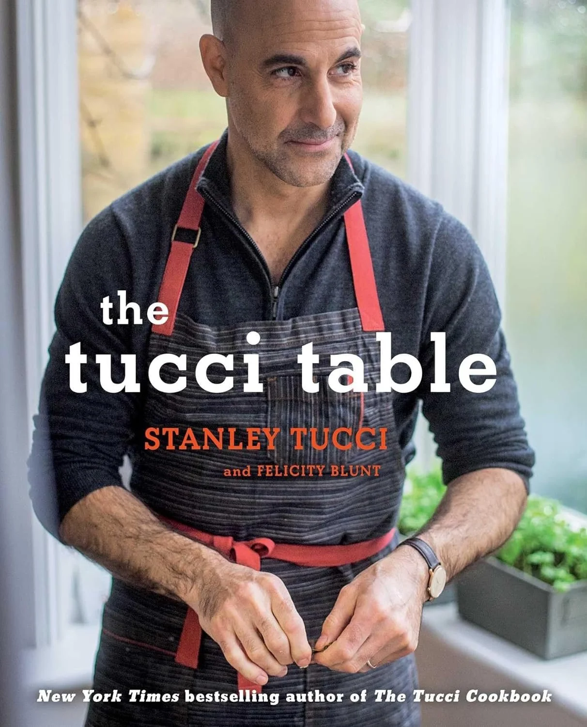Close-up of a man wearing a dark zip-up sweater and a striped apron with red straps, standing near a window with greenery outside. The book cover titled 'the tucci table' by Stanley Tucci and Felicity Blunt is overlayed with additional text about Tucci being a New York Times bestselling author.