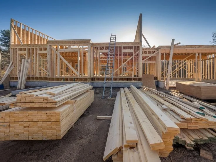 Construction site with wooden framework for a house in progress, piles of lumber in the foreground, and a clear sky.
