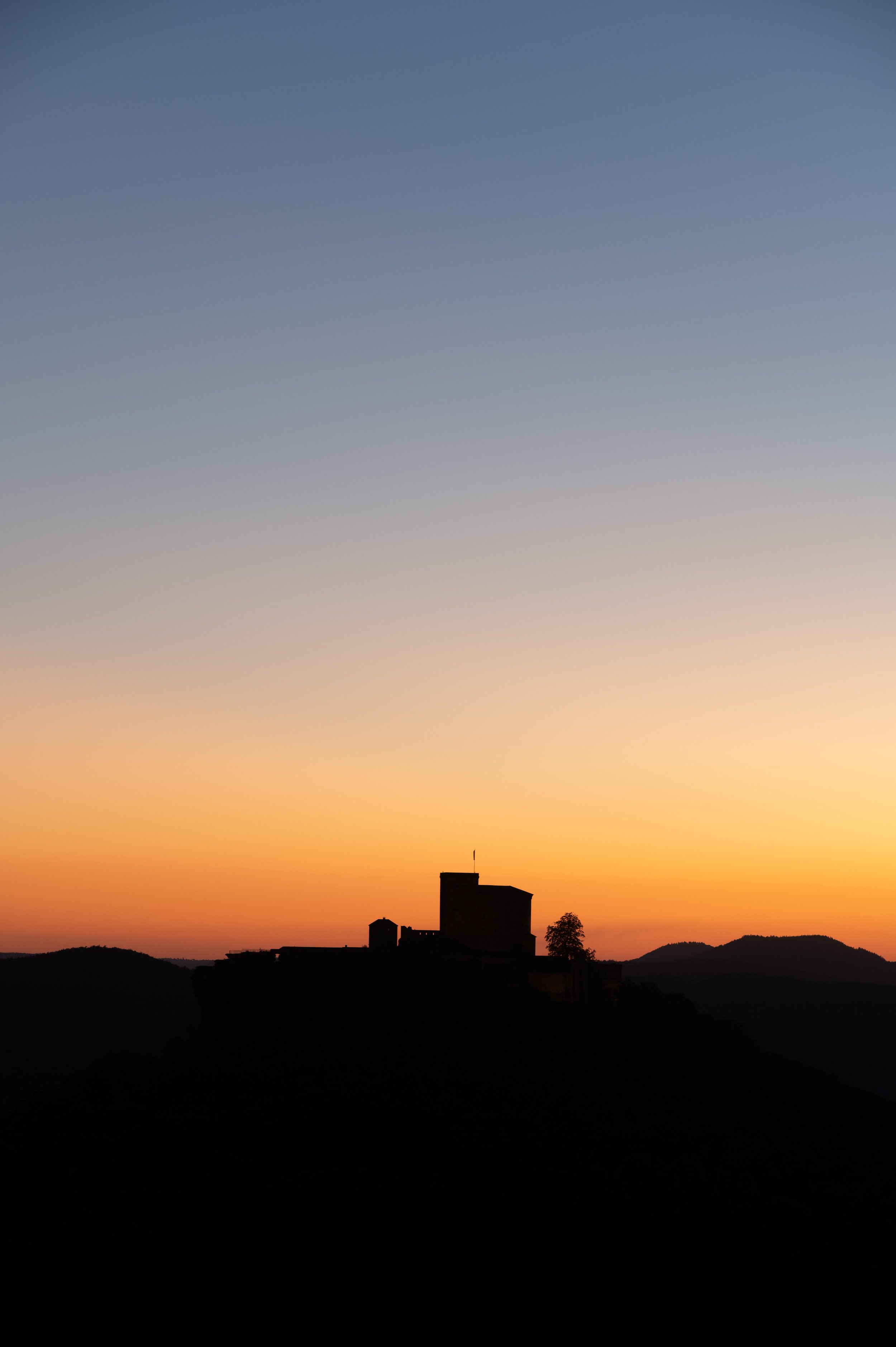 Silhouette einer Burg auf einem Hügel vor einem Sonnenuntergangshimmel mit orangefarbenen und blauen Tönen.