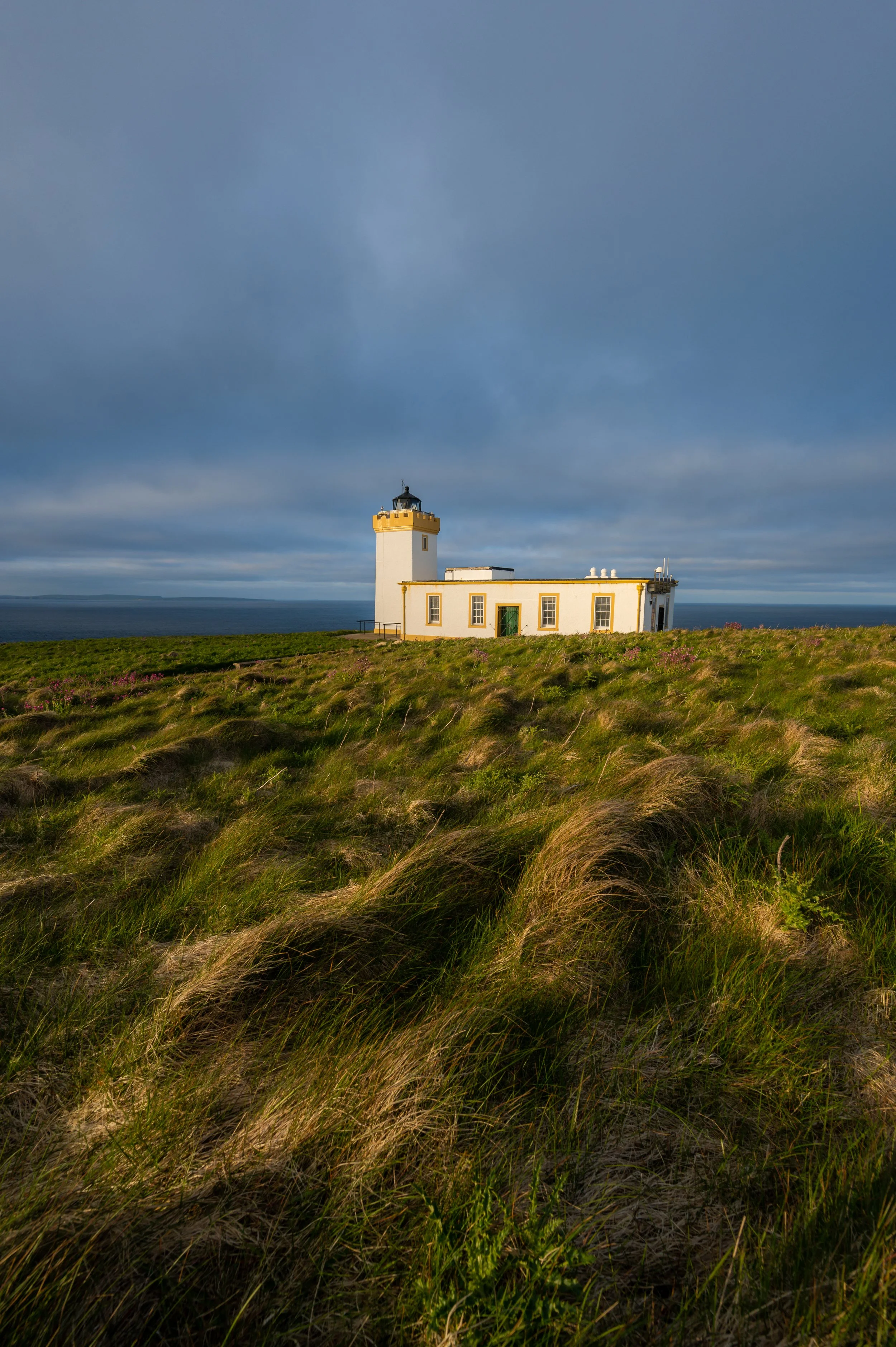 Leuchtturm auf einer grünen Wiese mit Blick auf das Meer, bewölkter Himmel im Hintergrund.