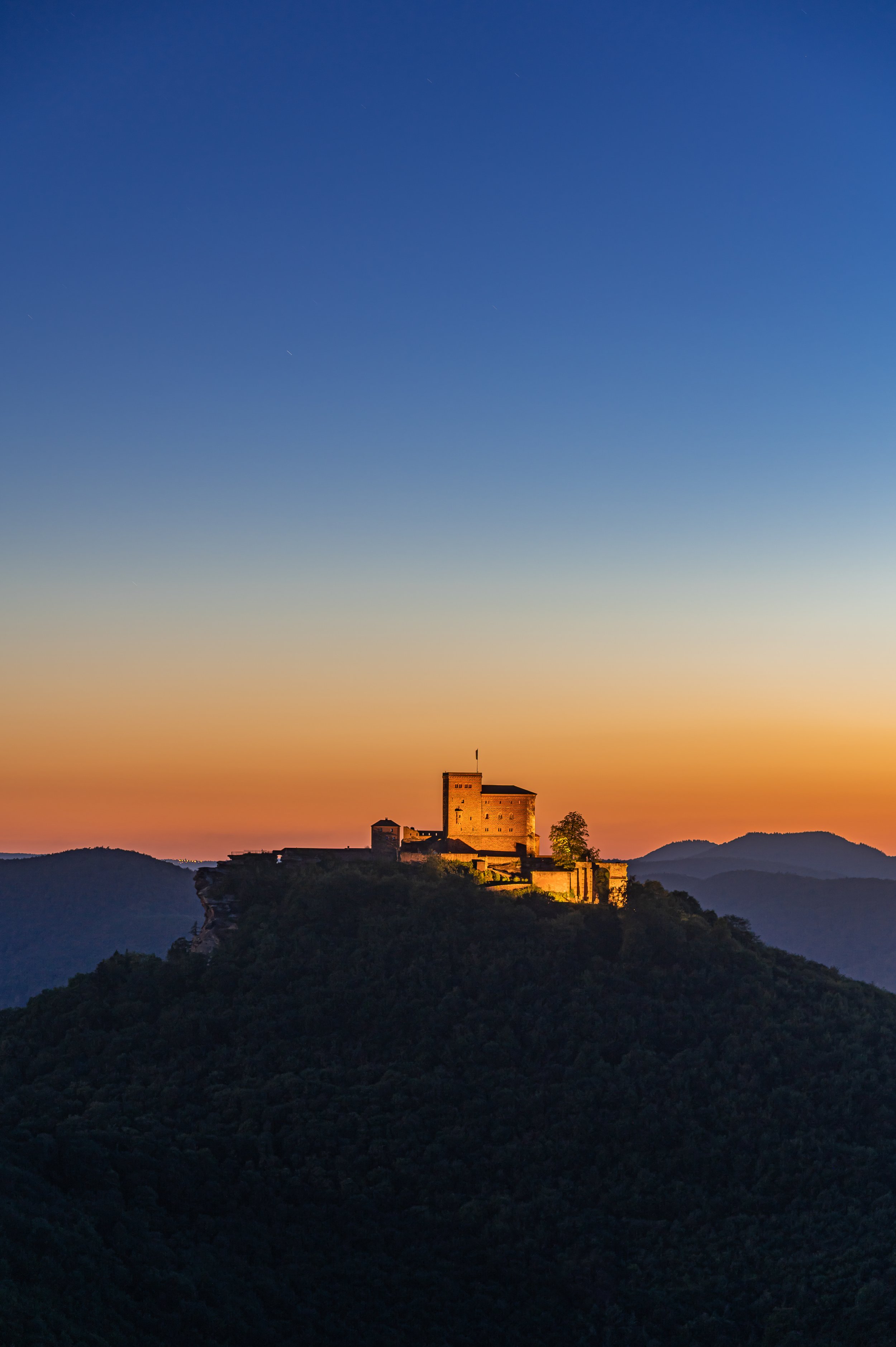 Burg auf einem Hügel bei Sonnenuntergang mit Bergen im Hintergrund und klarem Himmel.