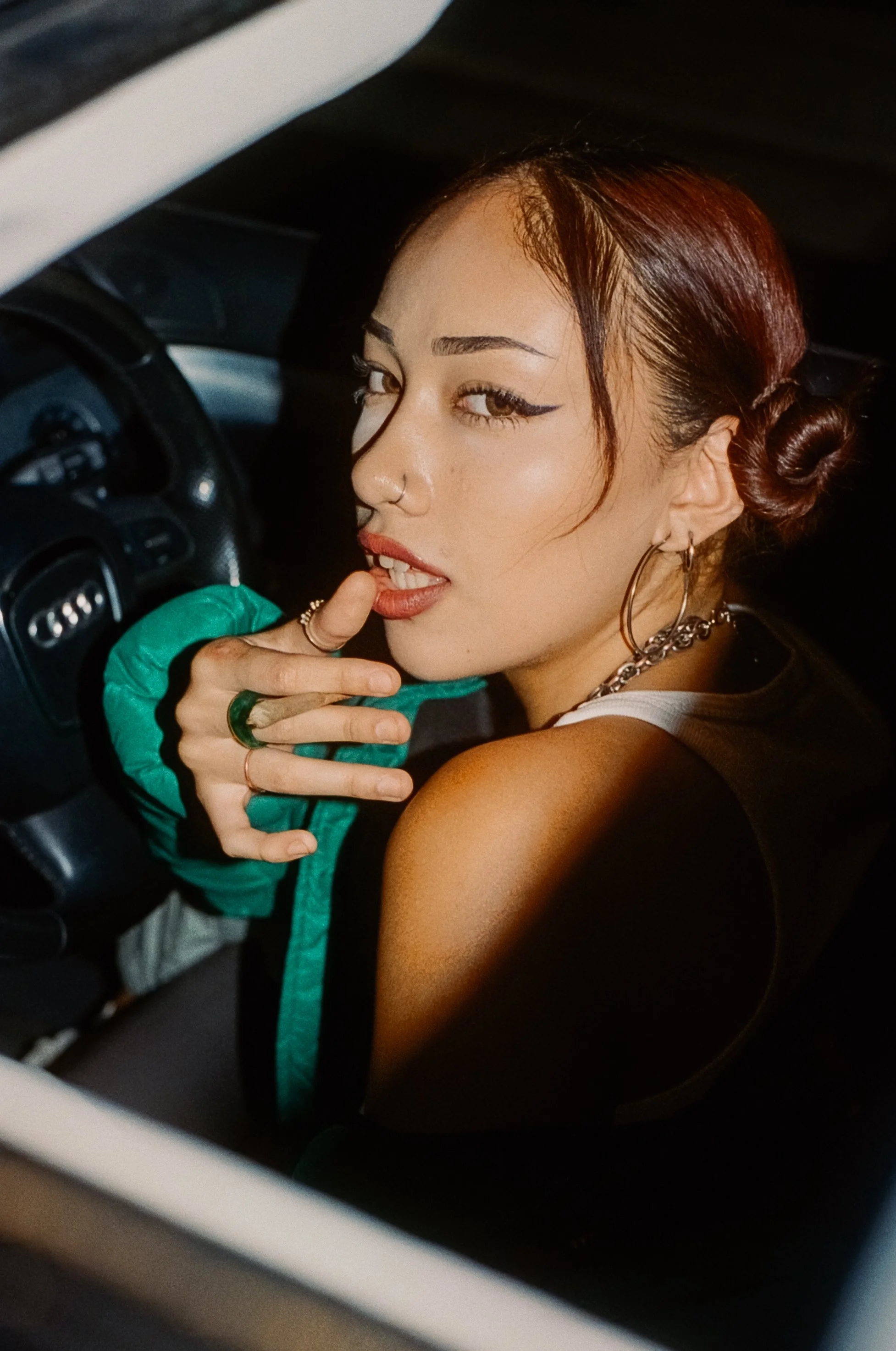 A woman with red hair styled in buns, wearing large hoop earrings and a chain necklace, looking over her shoulder inside a car.