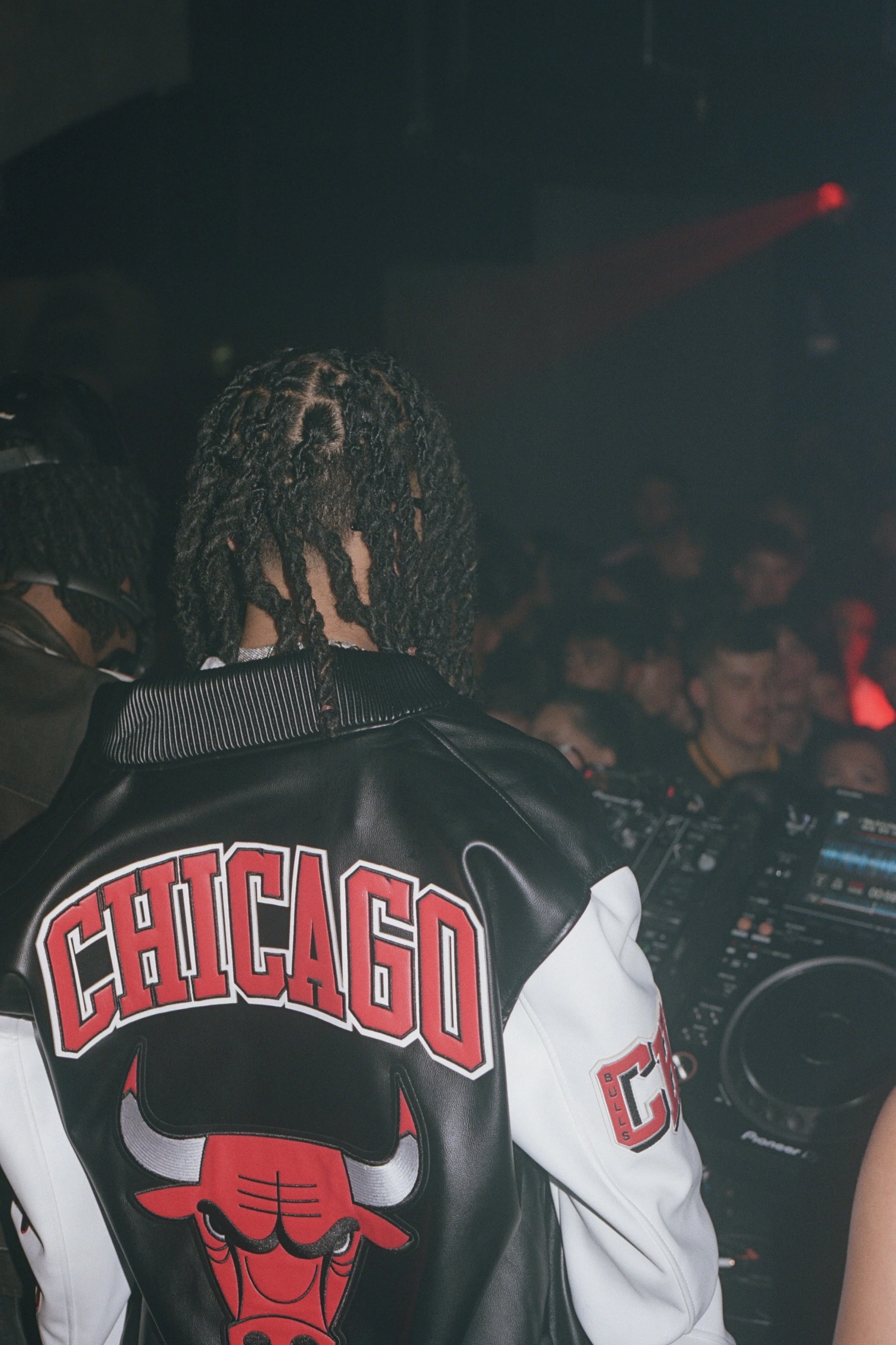 Person with dreadlocks wearing a black and white Chicago Bulls jacket, standing in front of DJ equipment at a nightclub or party.