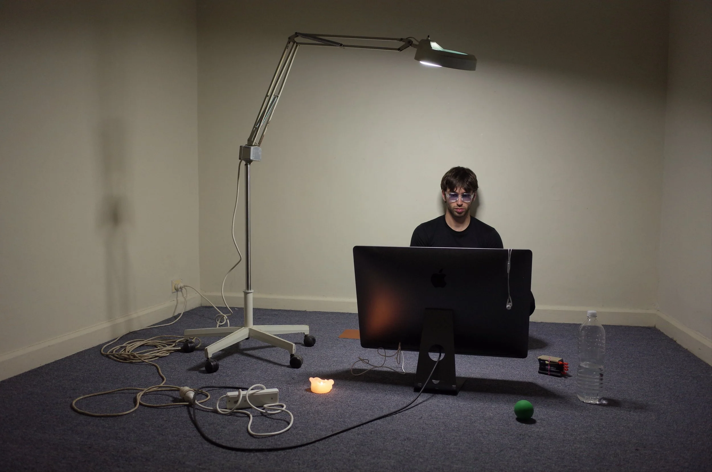 A young man with sunglasses sitting in a plain room at a desk with a computer, surrounded by clutter including a floor lamp, cables, a candle, a water bottle, and a green ball.