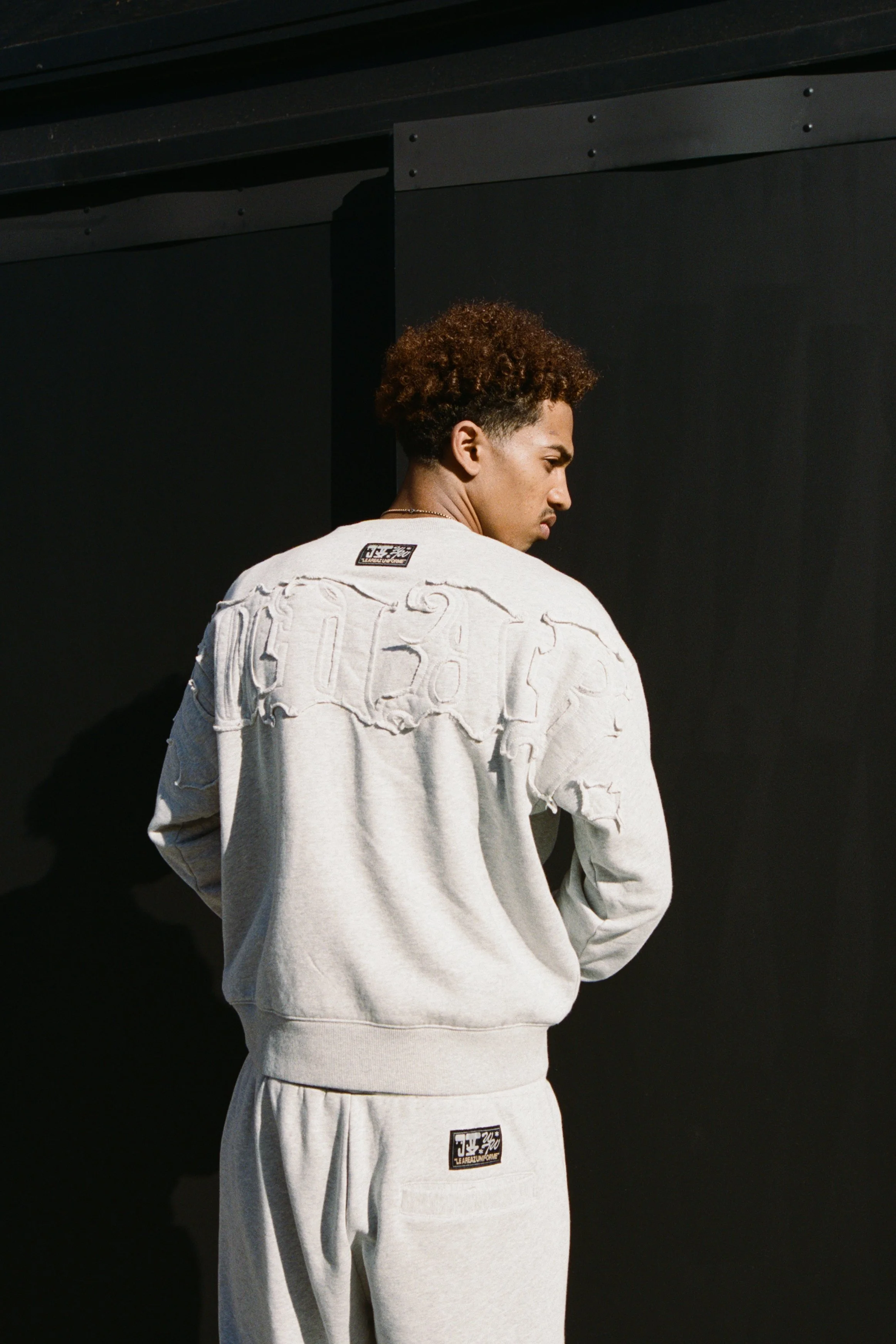 A young man with curly hair wearing a distressed gray sweatshirt and sweatpants, standing against a black background.