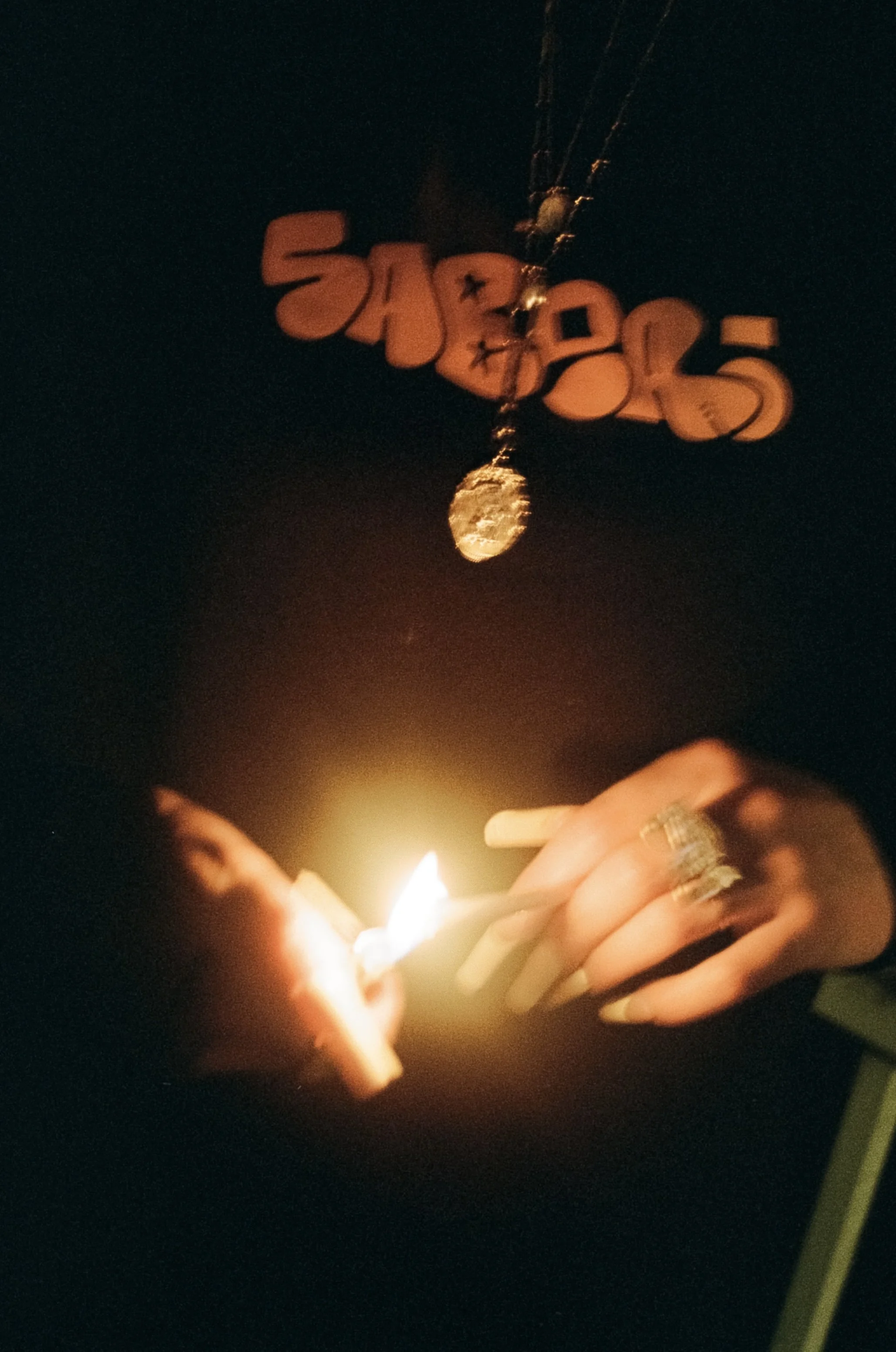 Close-up of hands holding lit matchstick and lighter, illuminating the dark surroundings. The person is wearing rings, including a large ring on the right hand, with a decorated nail on the left ring finger. In the background, hanging from the ceilin