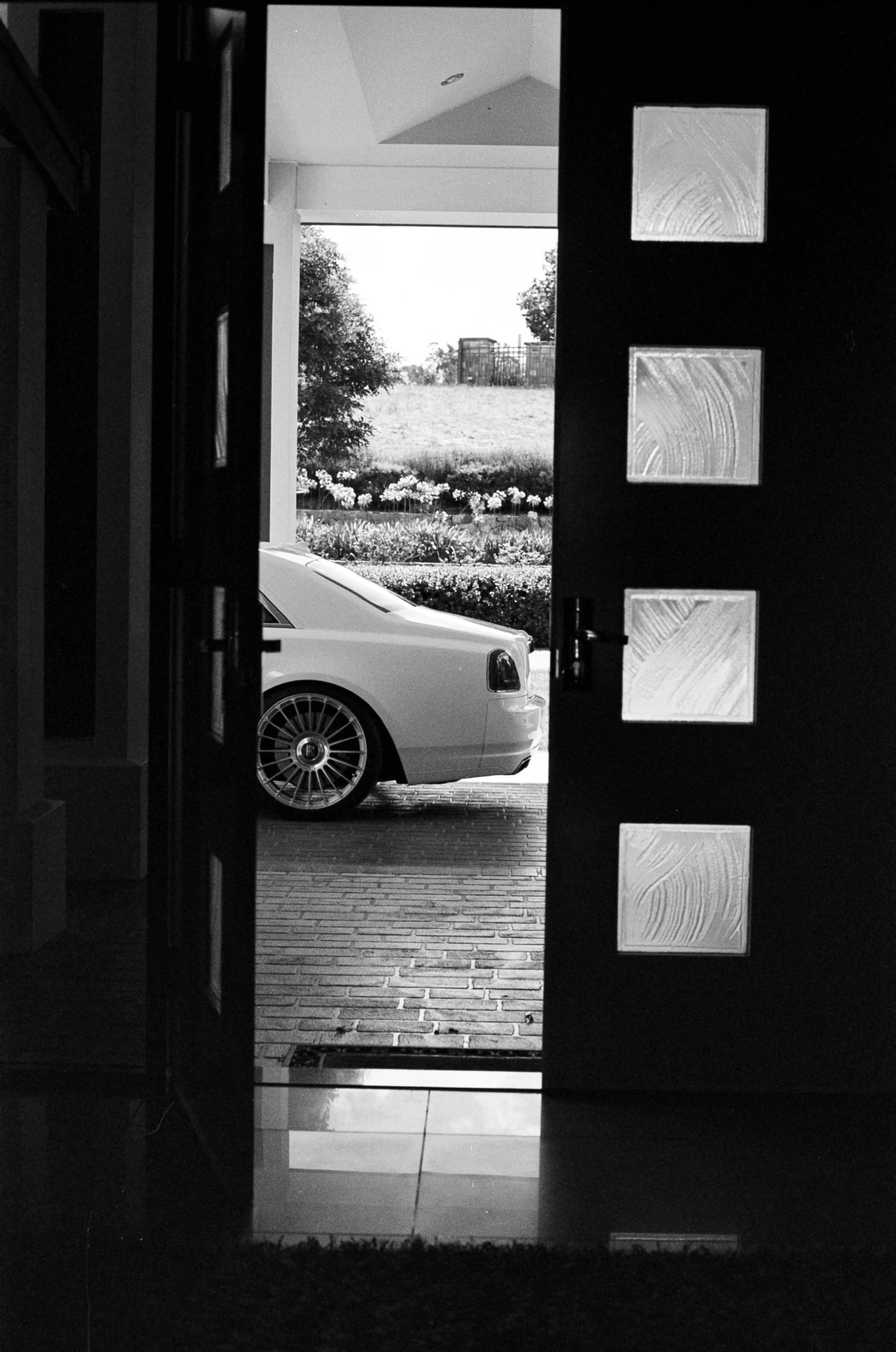 Black and white photo of an open front door viewed from inside, revealing a car parked in the driveway and a garden with flowers and trees outside.