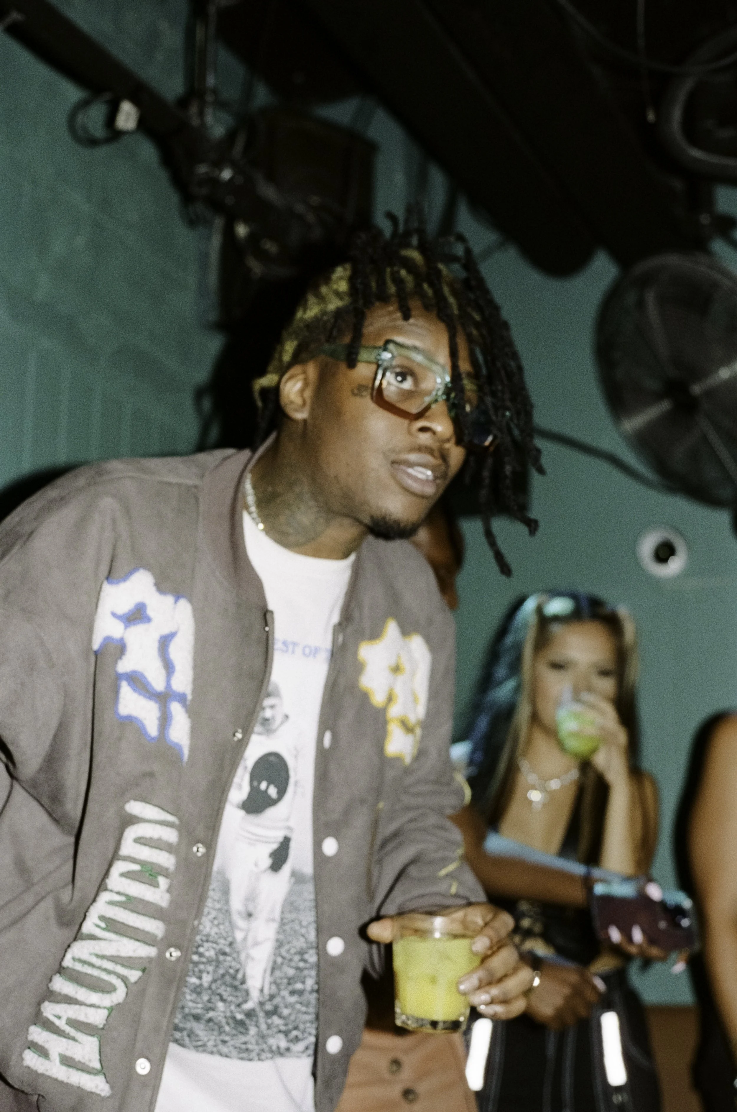 A young man with dreadlocks and glasses holding a yellow drink in a glass, standing in a room with teal walls, with two young women in the background, one holding a drink and the other holding a smartphone.