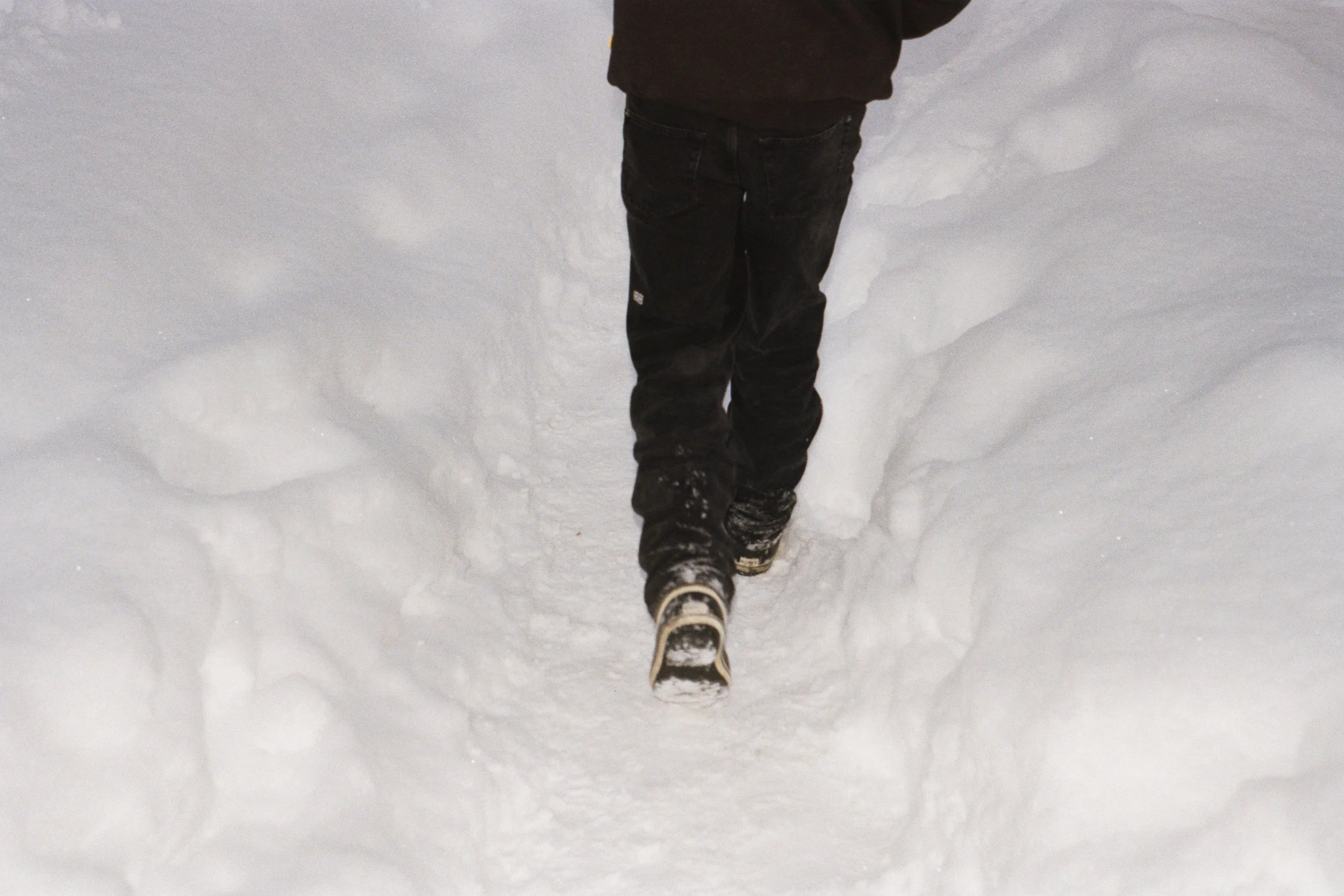 Person walking on snow-covered ground, wearing dark pants and shoes, leaving footprints.