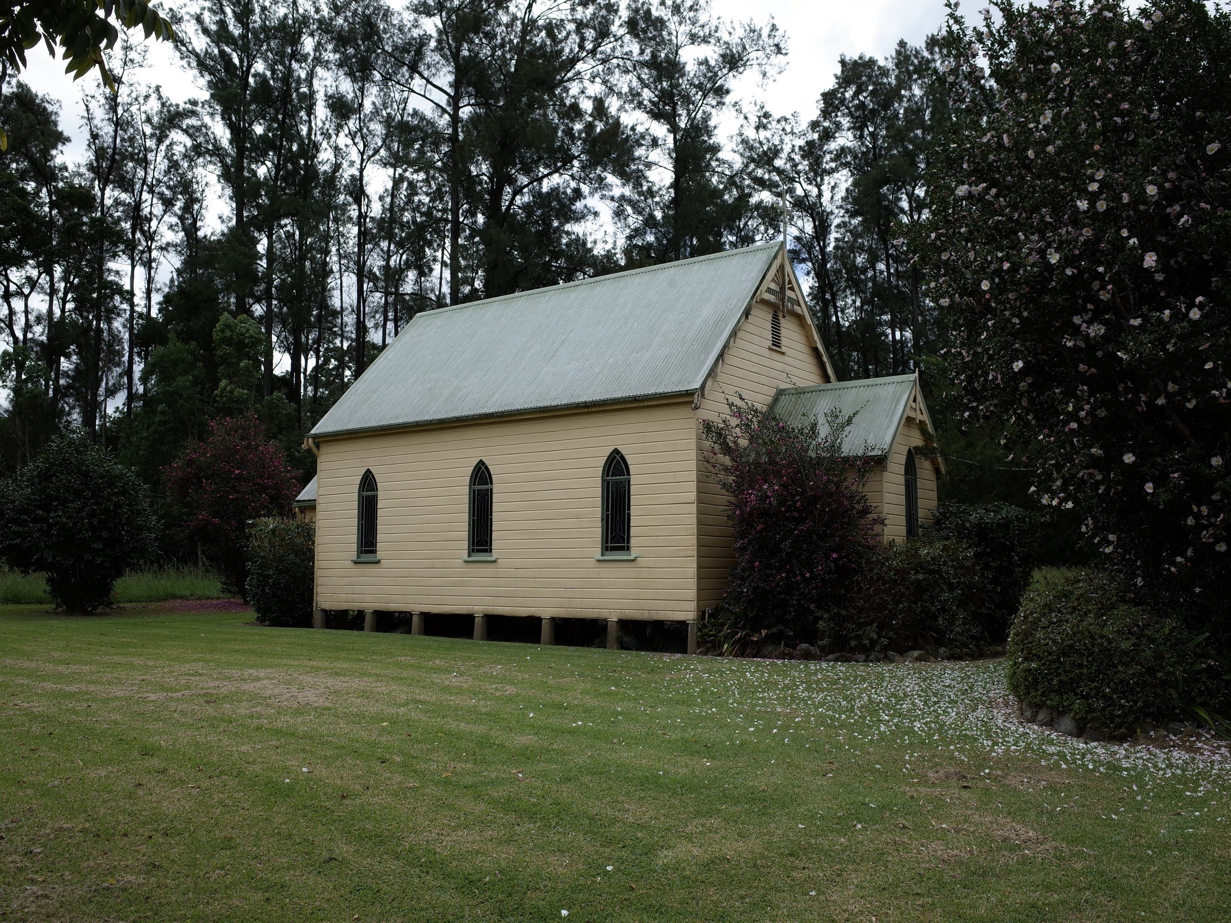 A small, yellow, wooden church with arched windows, surrounded by green grass, flowering bushes, and dark green trees in the background under a cloudy sky.