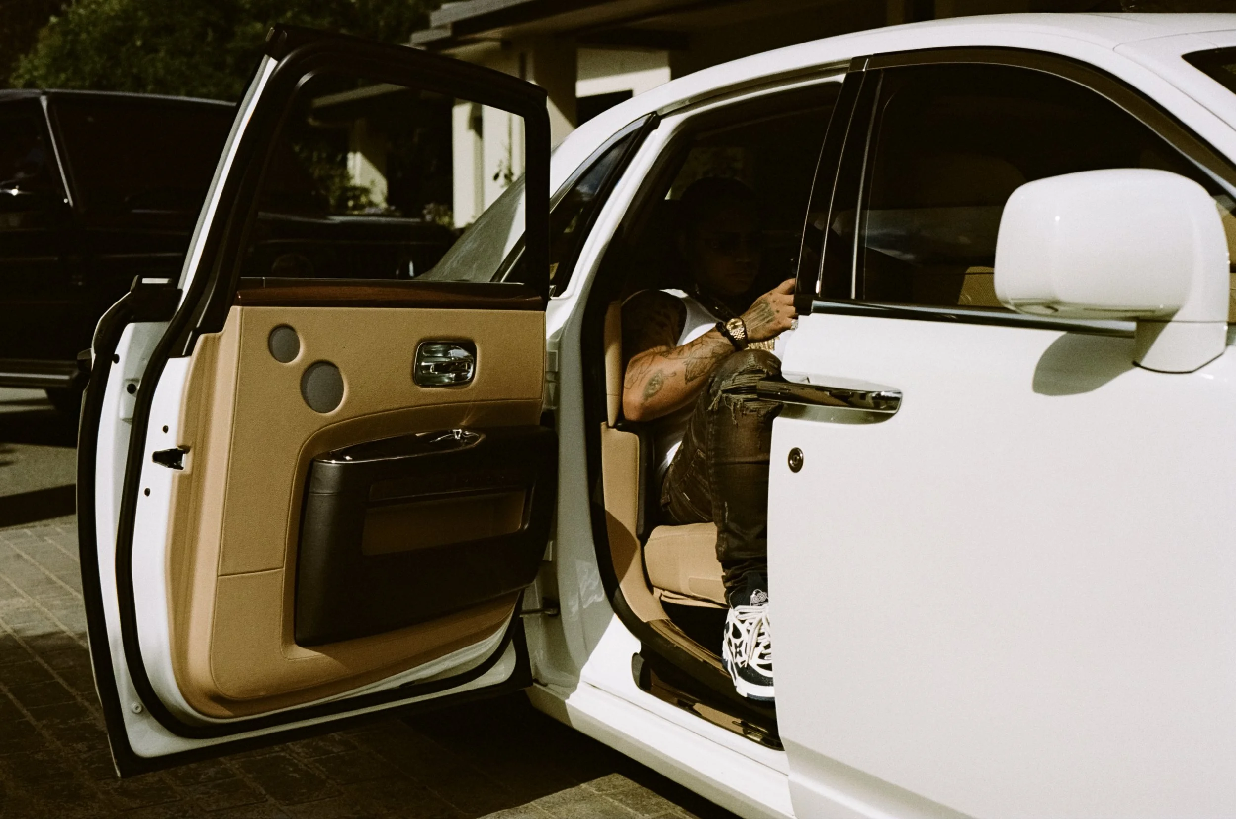 A person with tattoos and wearing a watch sitting in a white luxury car with tan interior, looking at their phone, with the car door open.