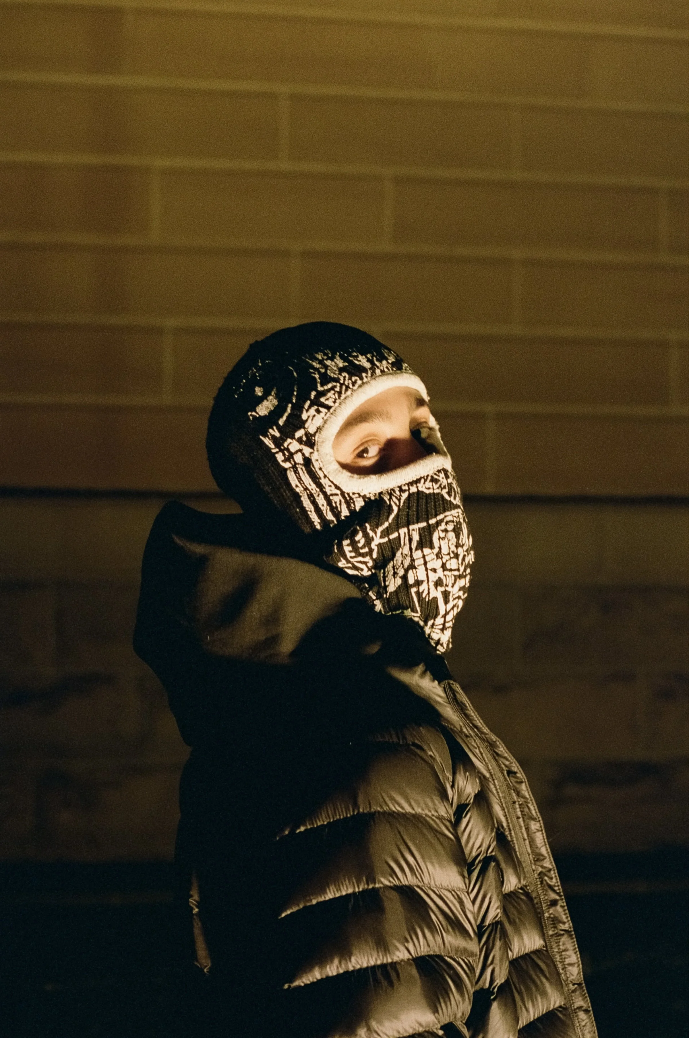 A person wearing a black and white patterned ski mask, black hoodie, and shiny black puffer jacket, standing outdoors at night with a brick wall in the background.