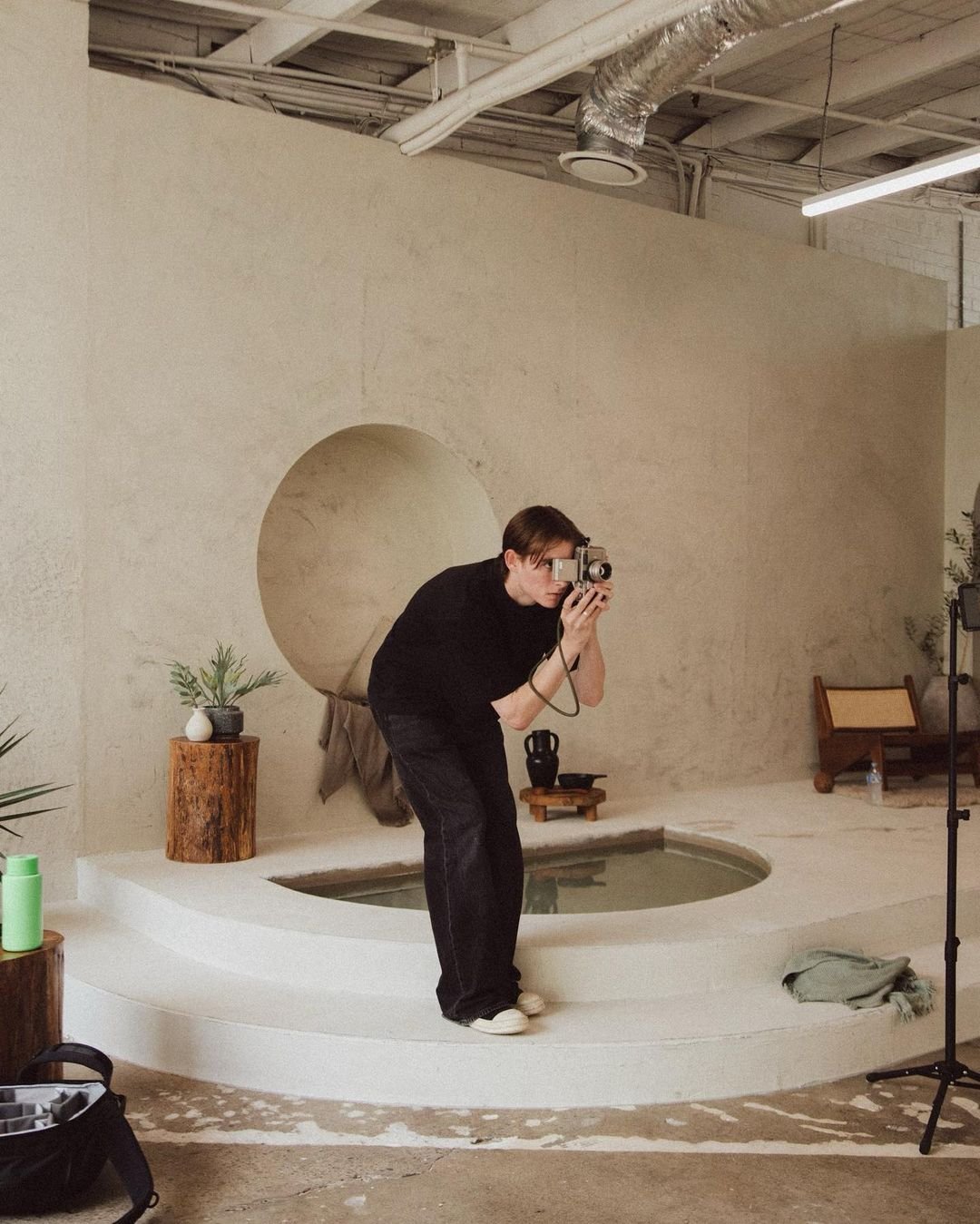 A person taking a photo of themselves in an art studio or gallery, standing on a small raised platform with a round cutout behind them, surrounded by minimalist decor including plants and wooden accessories.