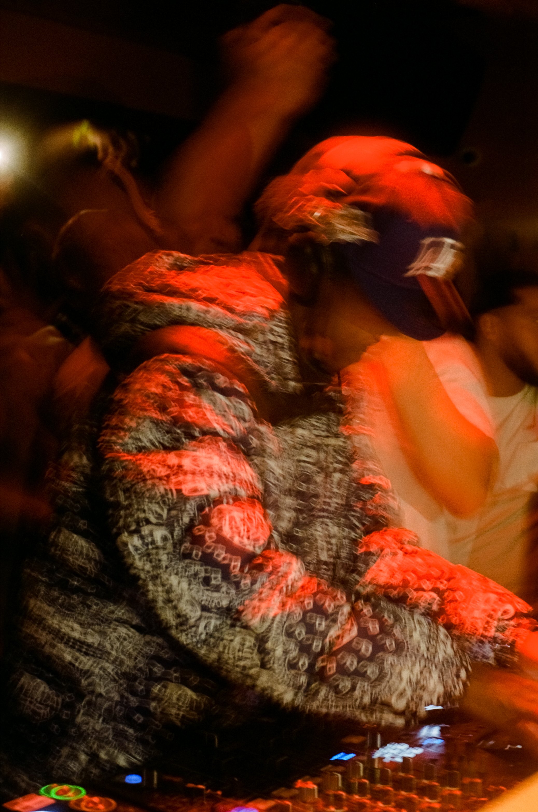 A DJ wearing a cap and patterned clothing, mixing music on a DJ console in a dimly lit setting with red and orange lights.