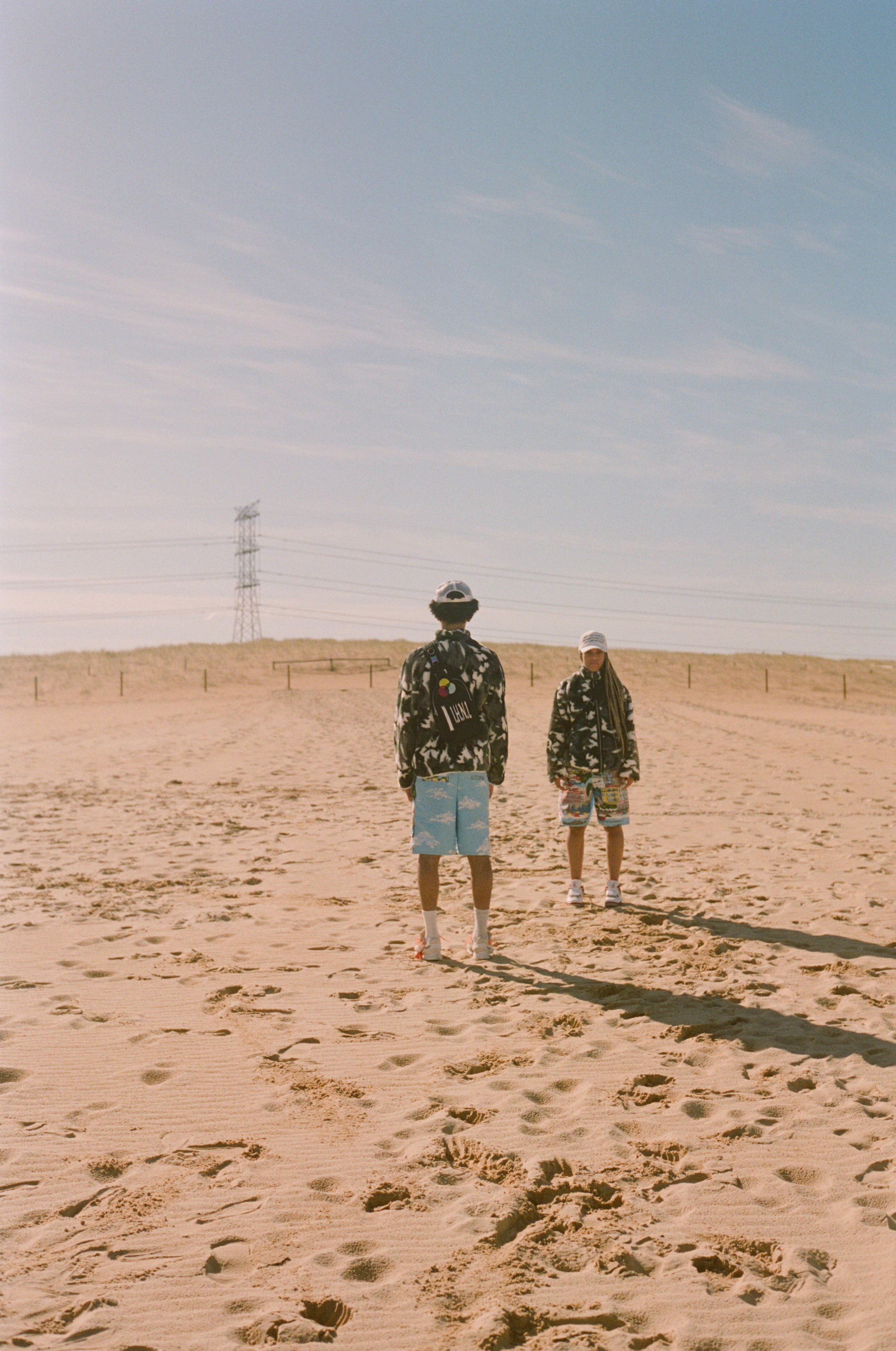 Two people standing on a sandy beach, facing each other, with one wearing a white cap and shorts, the other with a backpack and a hat, under a light blue sky with wispy clouds, power lines, and a transmission tower in the background.