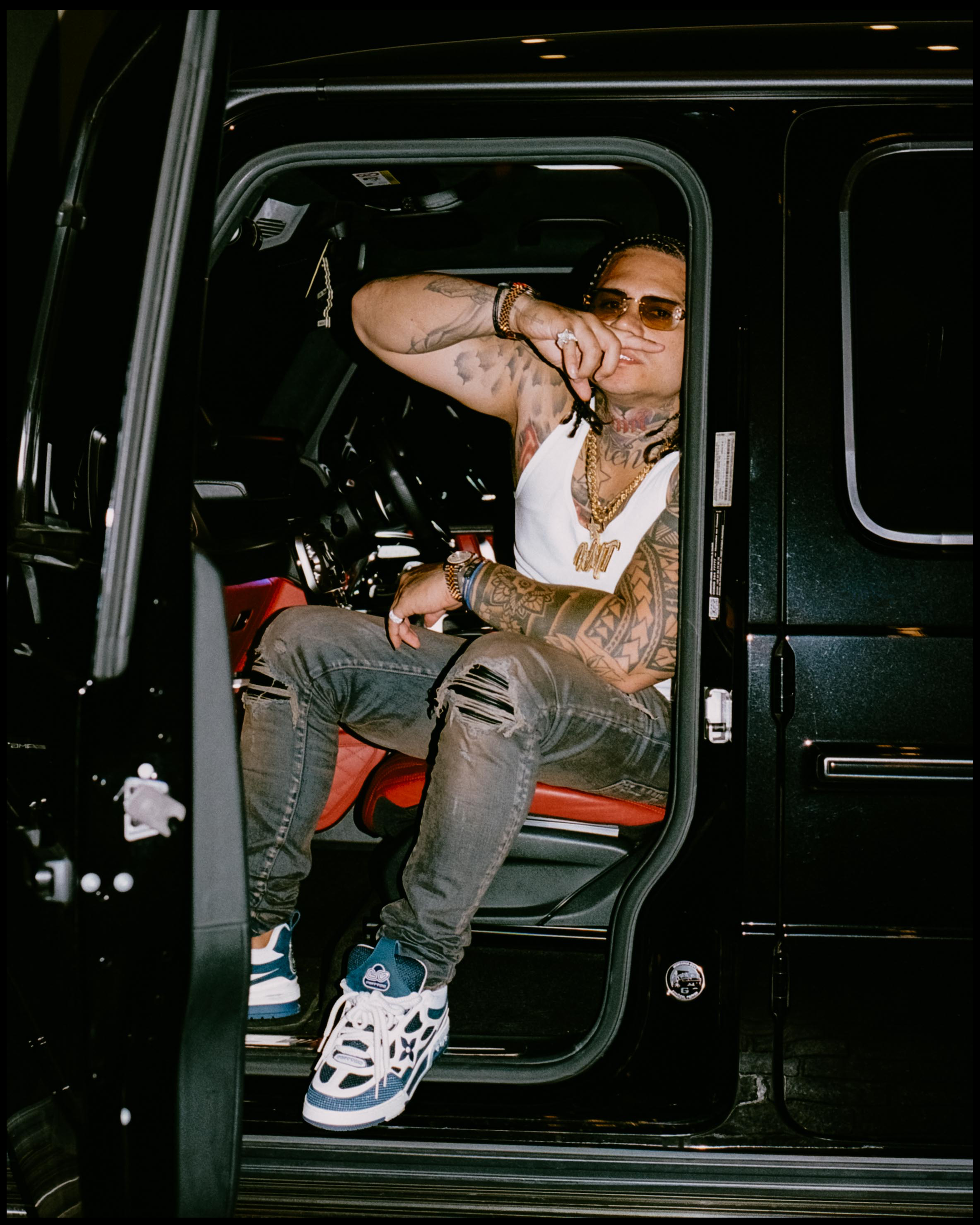 A young man with tattoos on his arms and neck, wearing glasses, jewelry, a white tank top, and distressed jeans, sitting in the driver's seat of a black vehicle. He is making a gesture with his right hand while looking out of the open window.