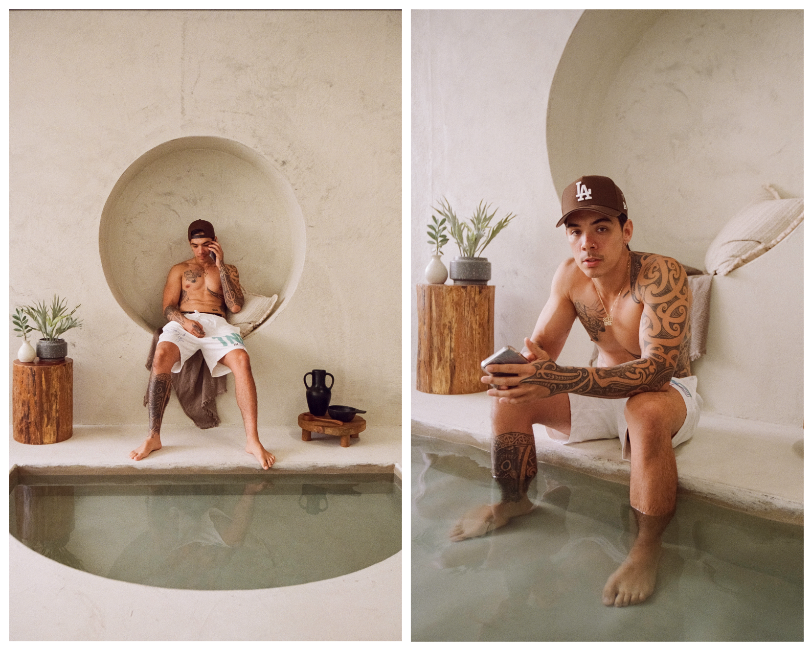 A young man with tattoos, wearing a baseball cap and white shorts, sitting with his feet in a pool, holding a phone and looking at the camera inside a minimalist room with a circular wall niche, wooden table, plants, and pottery.