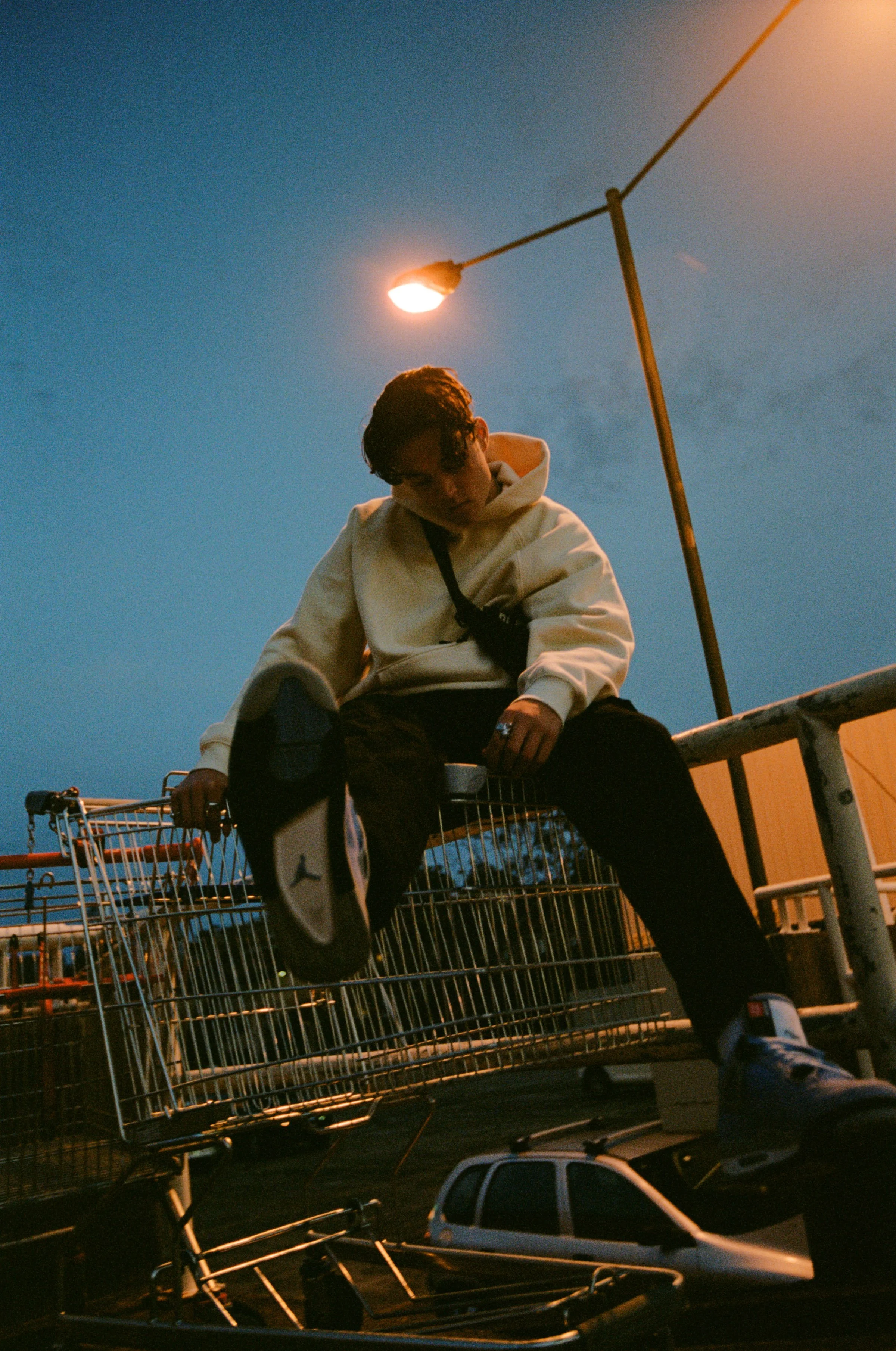 A young man with dark hair, wearing a white hoodie and dark pants, sitting on a car shopping cart in a parking lot during dusk. There are shopping carts and cars nearby, and a streetlamp is lit against a night sky.