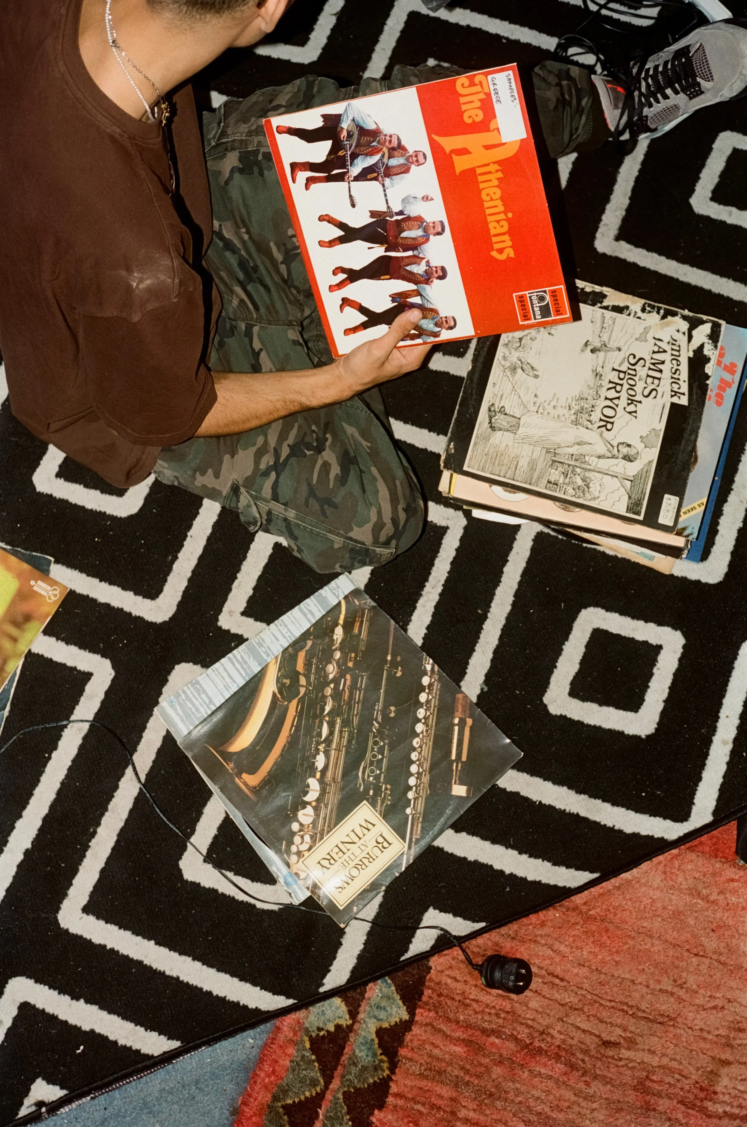 A person sitting on a black and white geometric patterned rug, holding a board game titled 'The Thieving)'. On the rug, there are other magazines or books, one with a saxophone and the title 'Bournemouth at the Winery'. The person is wearing a brown 