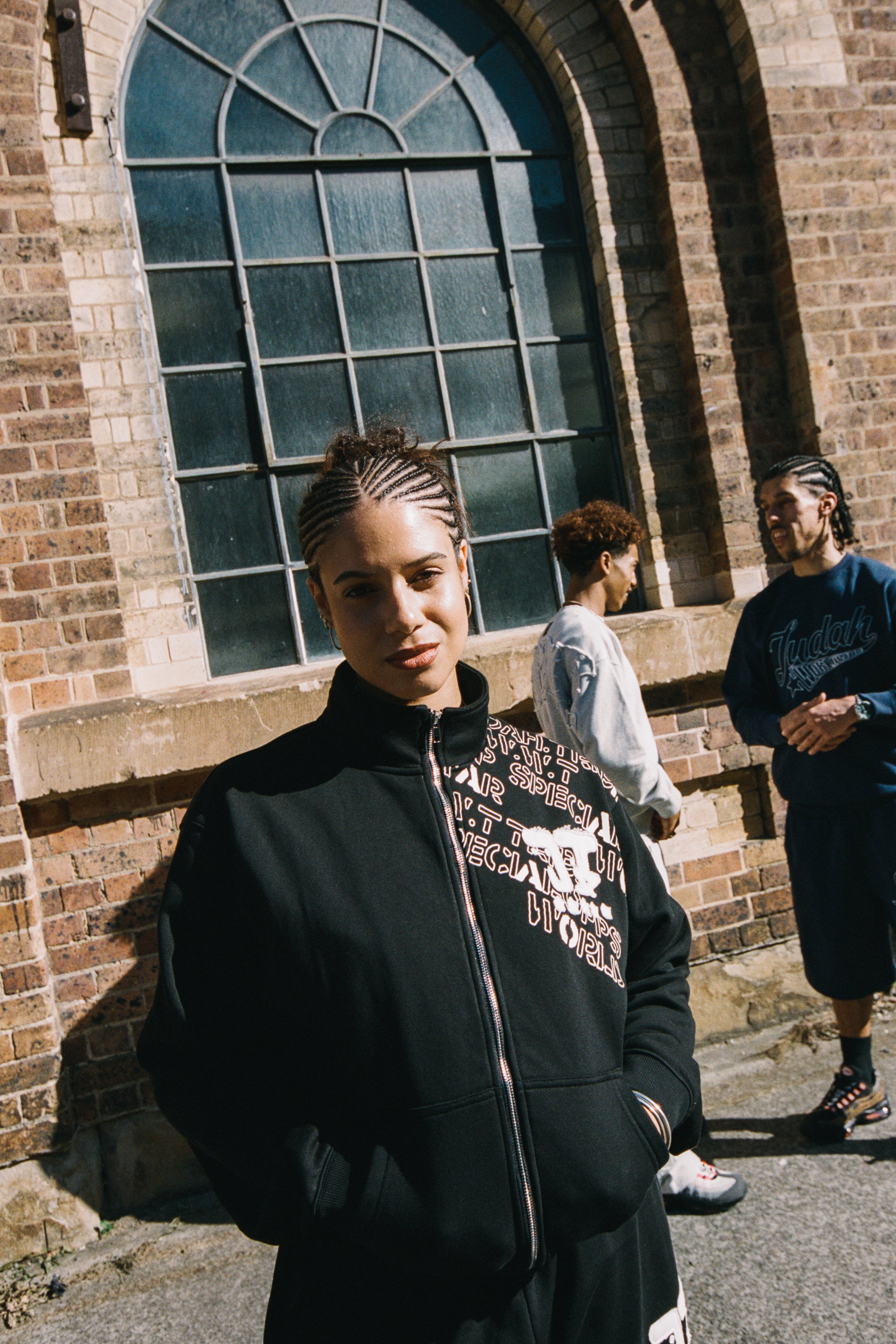 A young woman with braided hair and a black jacket standing in front of a brick building with large window, while two other people in casual clothing are in the background, smiling and talking.