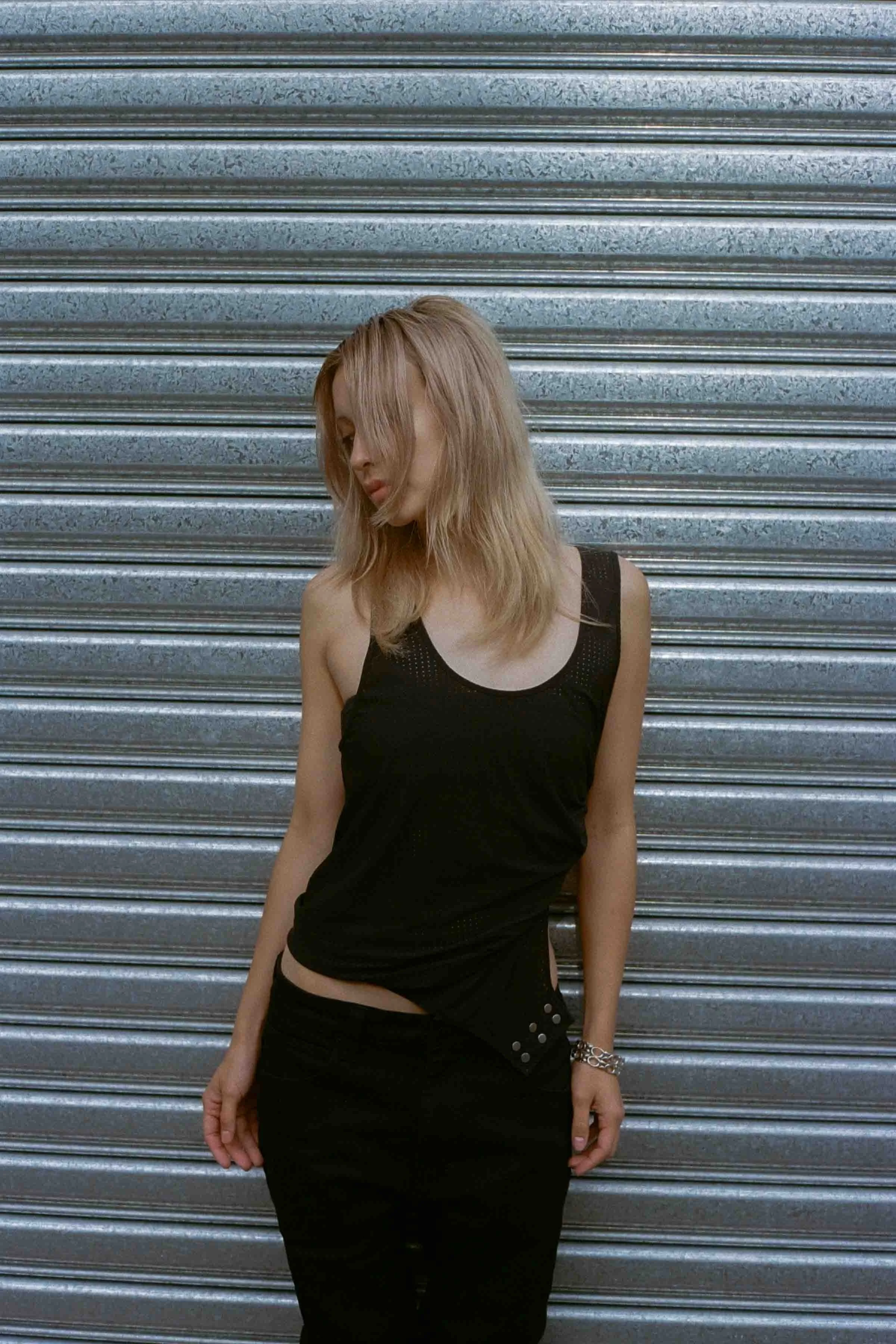 A young woman with blonde hair wearing a black sleeveless dress, standing against a metal garage door. She has her head tilted slightly downward, with partial hair covering her face.