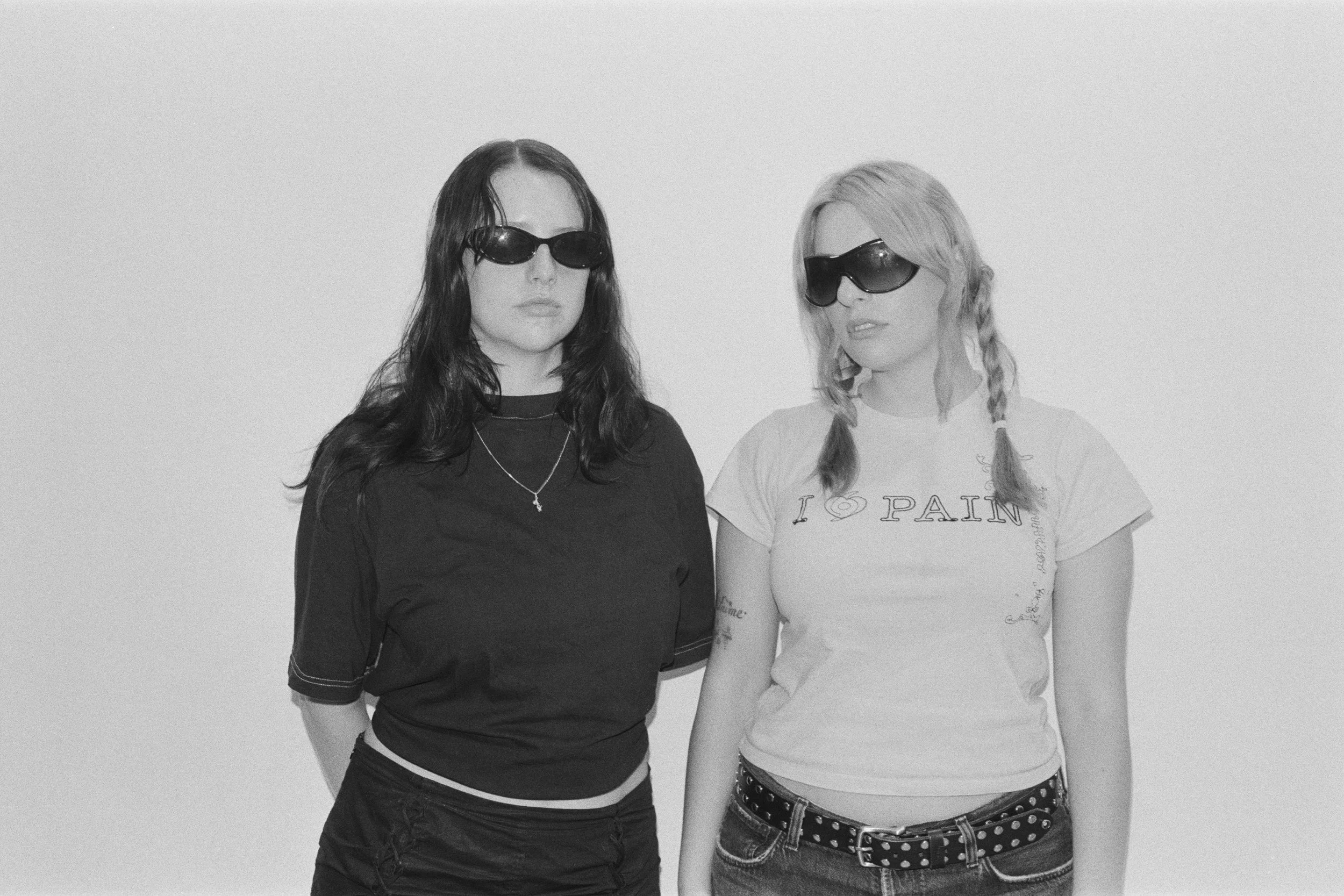 Two young women standing side by side against a plain background, both wearing large sunglasses and casual clothing.